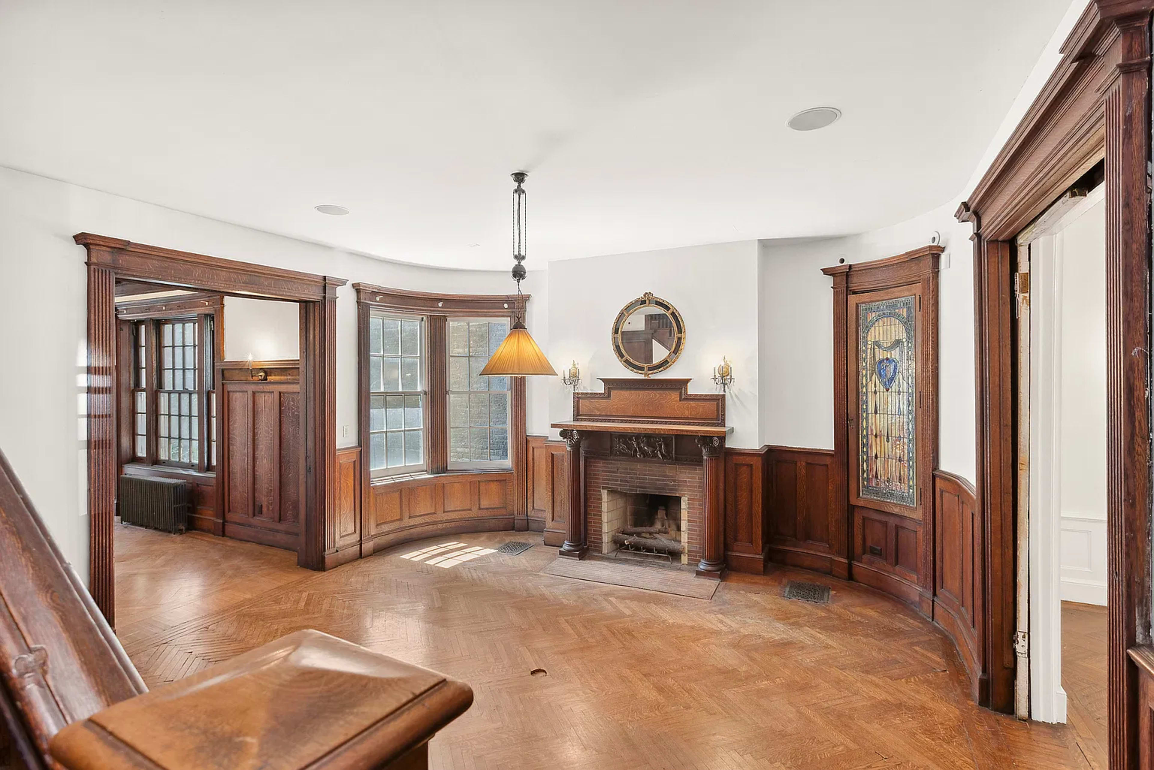 The image shows a spacious room with wooden paneling, a fireplace in the center, and large windows allowing natural light to enter.