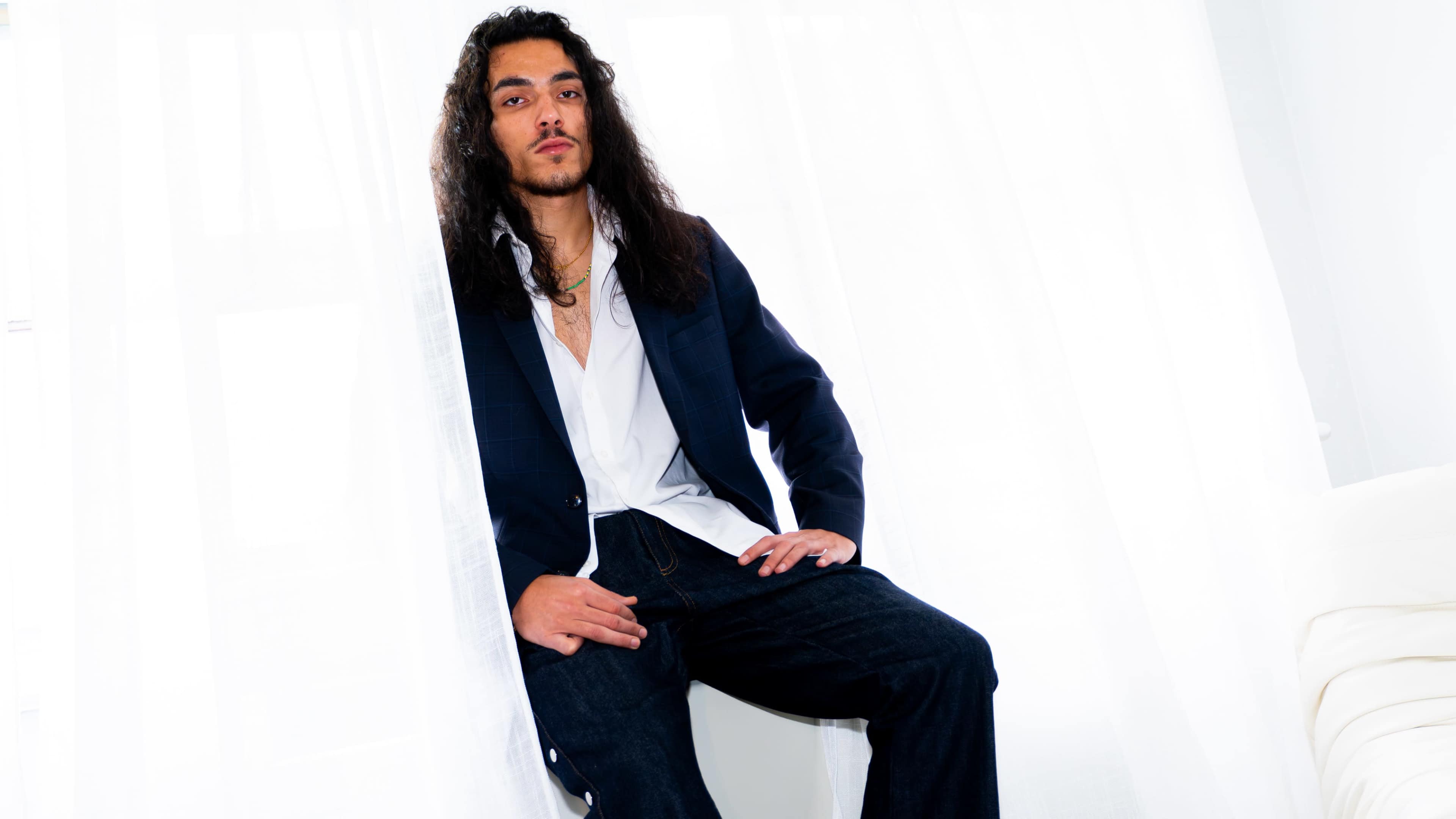 A man with long, wavy hair is sitting against a white curtain, wearing a blazer over a white shirt and dark jeans.