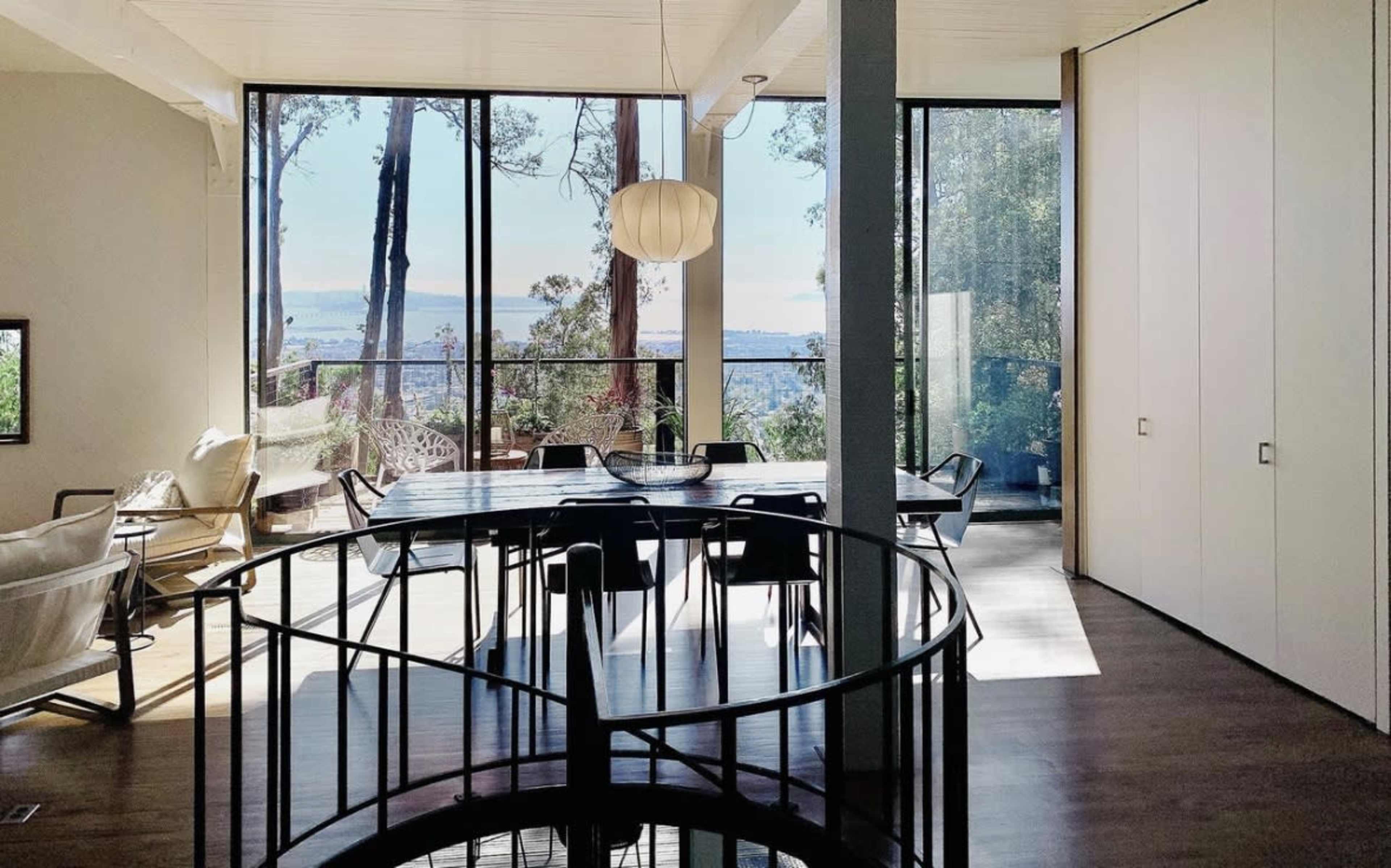 A spacious room features a large dining table surrounded by chairs, with large windows offering a view of trees and distant water.