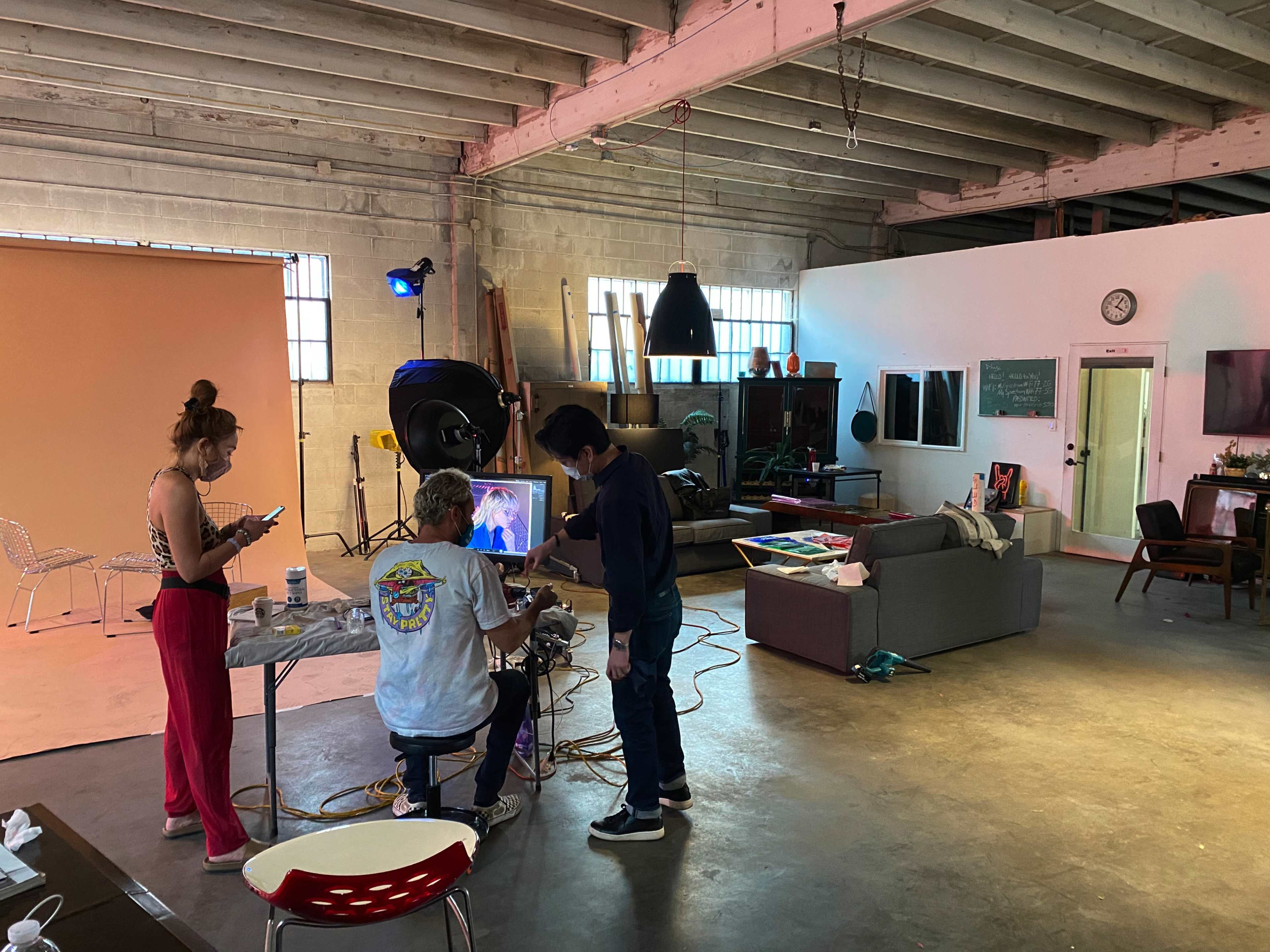 A studio space is set up for a photoshoot, featuring a model preparing in a chair, while a photographer adjusts equipment and another person uses a phone nearby.