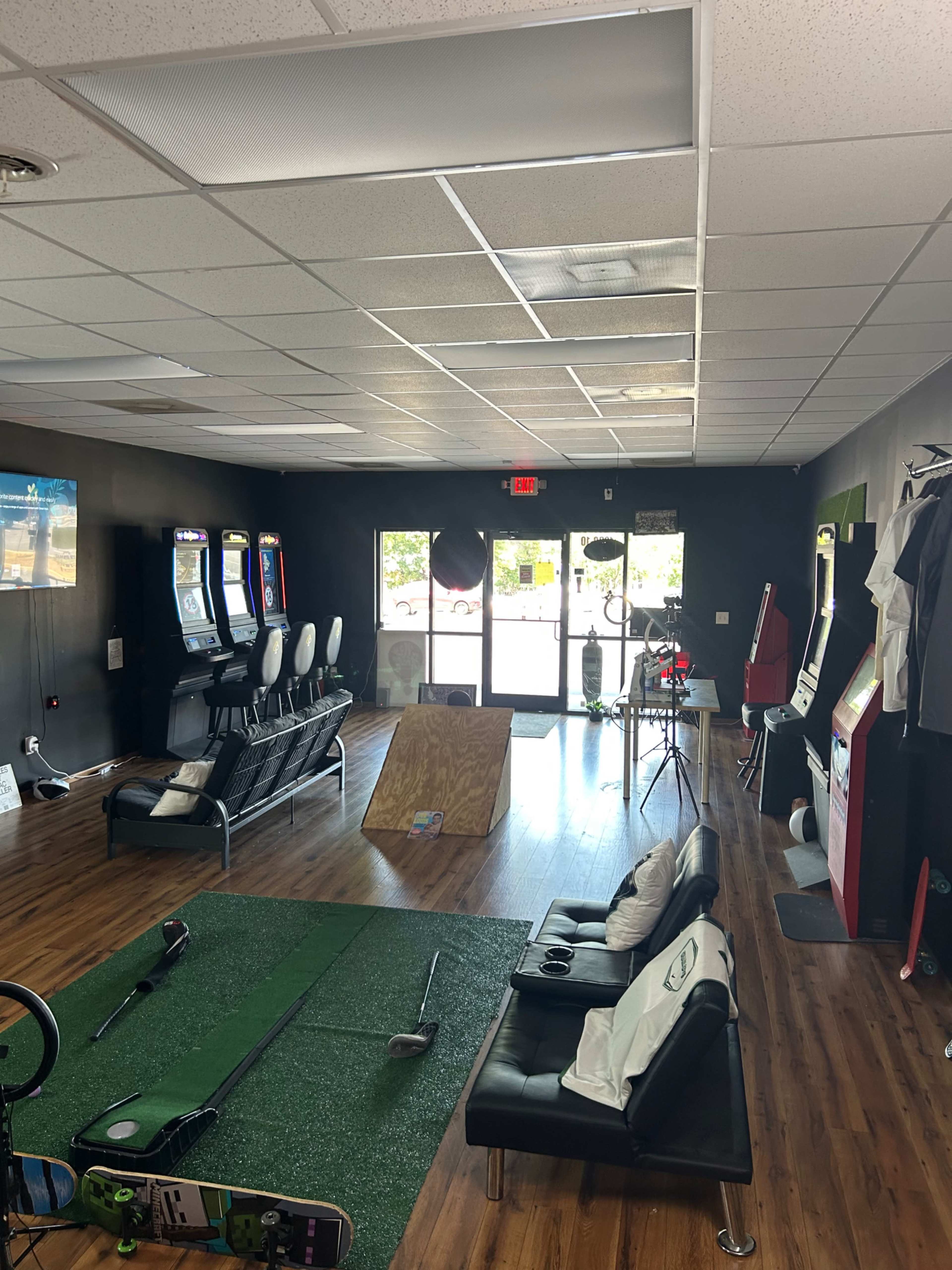 The image shows an indoor recreational space with arcade machines, a putting green, a wooden ramp, and seating arranged along a wooden floor.