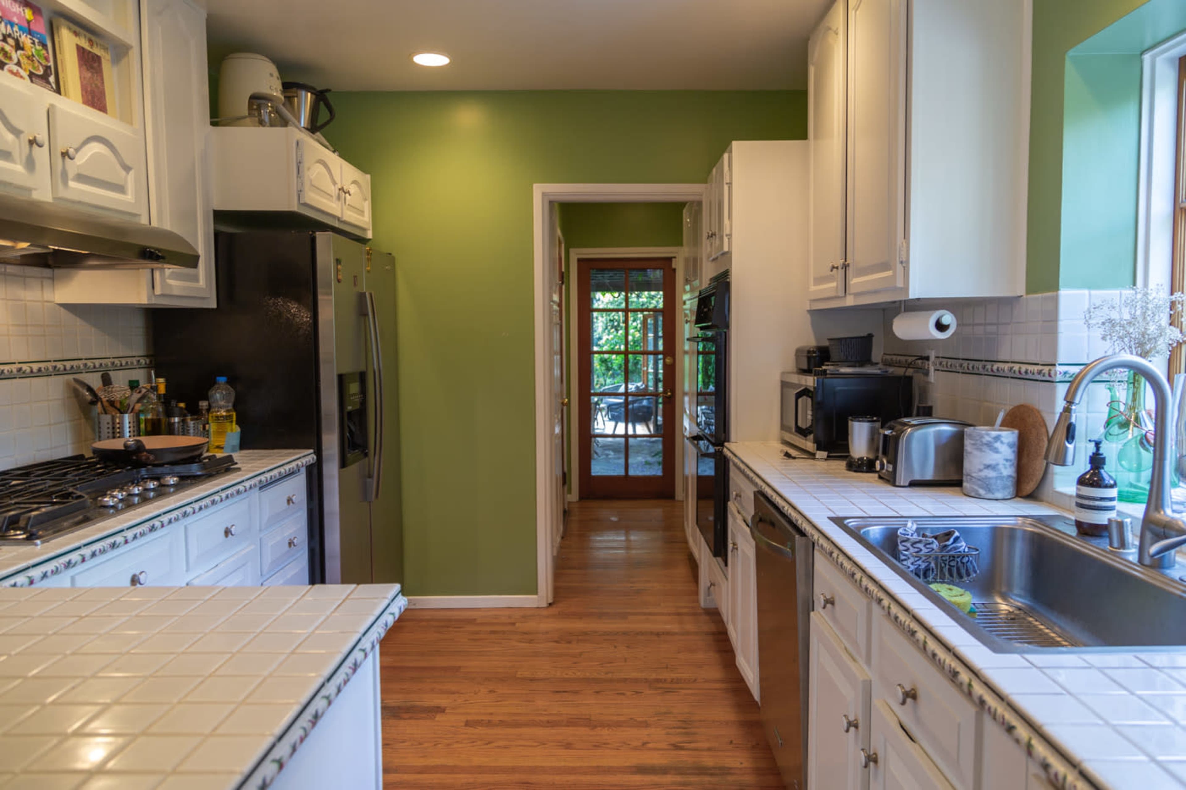 A spacious kitchen features white cabinets and countertops with green walls, leading to a doorway that opens to an outdoor area.