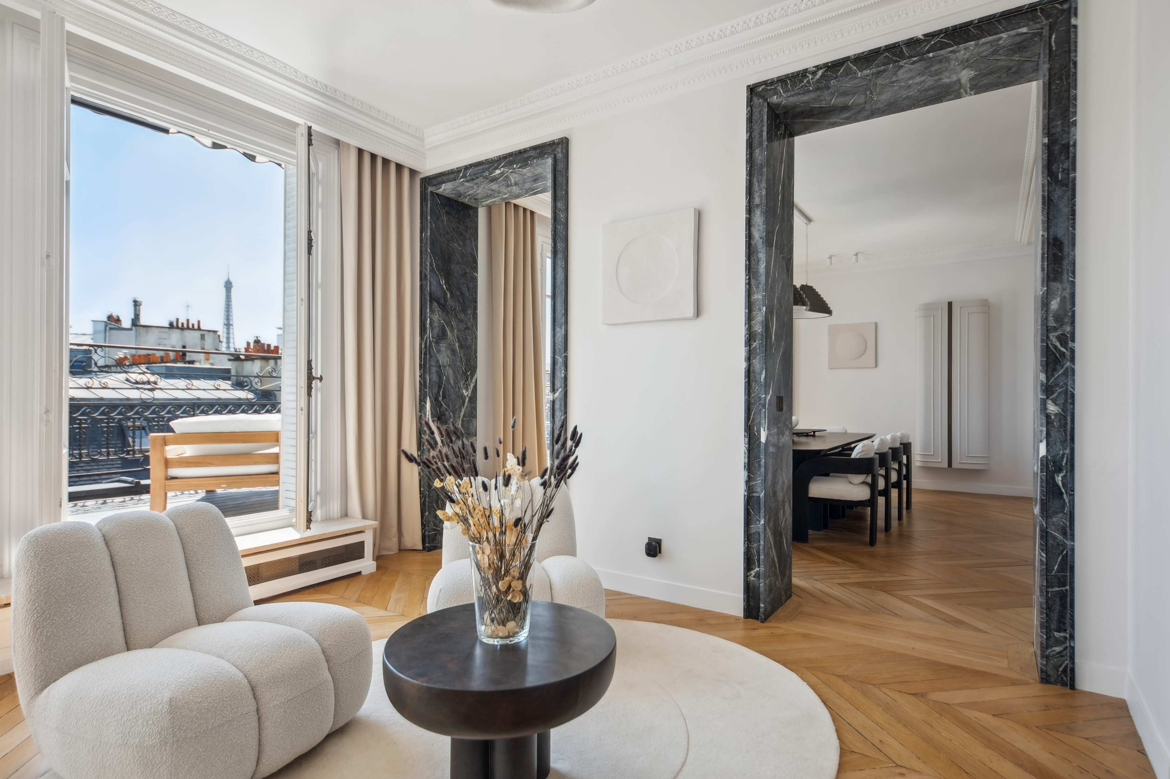 A modern living room with a round table and a plush armchair, featuring a large window that offers a view of rooftops and the Eiffel Tower in the background.