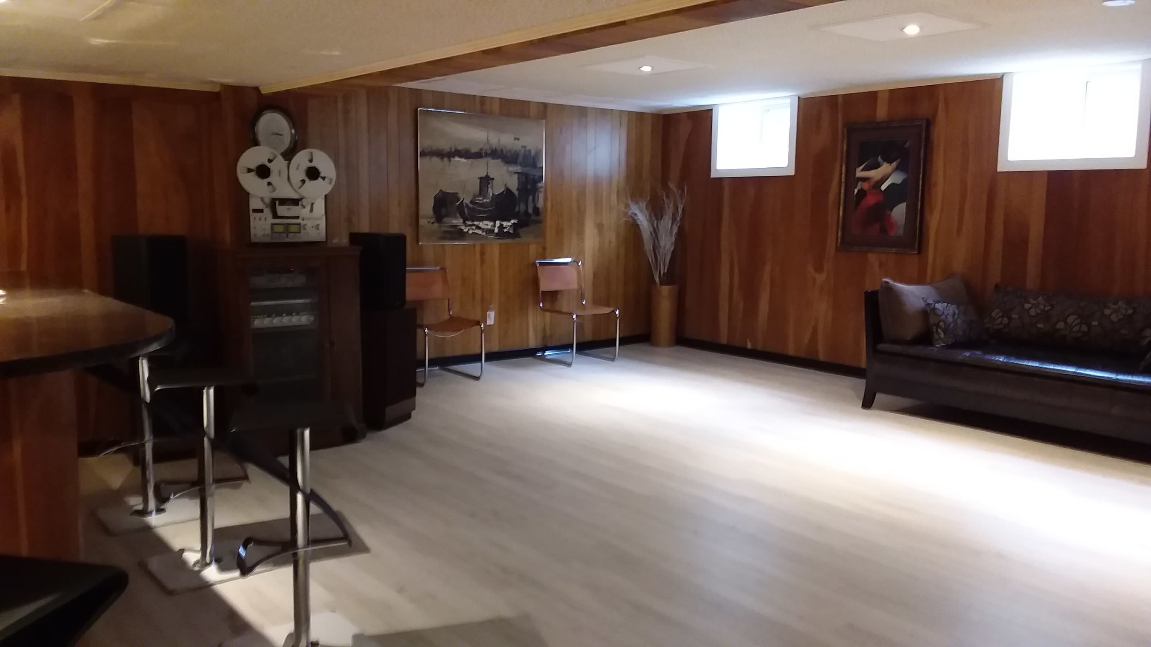 A spacious basement room with wooden paneling, featuring a seating area with a dark couch, two chairs, a bar counter, and a vintage projector.