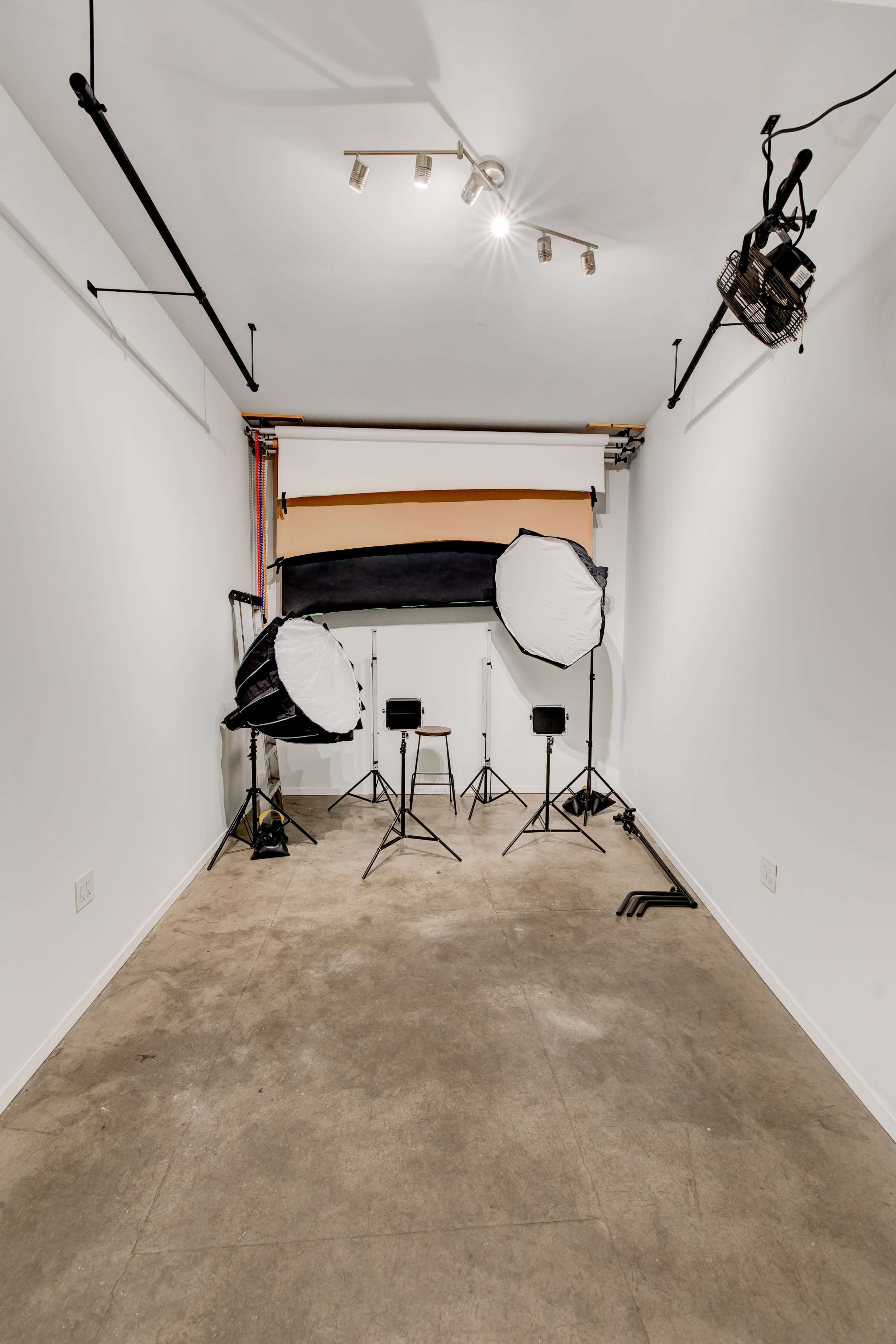 The image shows a photography studio with a plain white backdrop, two light stands with softboxes, and two chairs positioned in front of the backdrop.