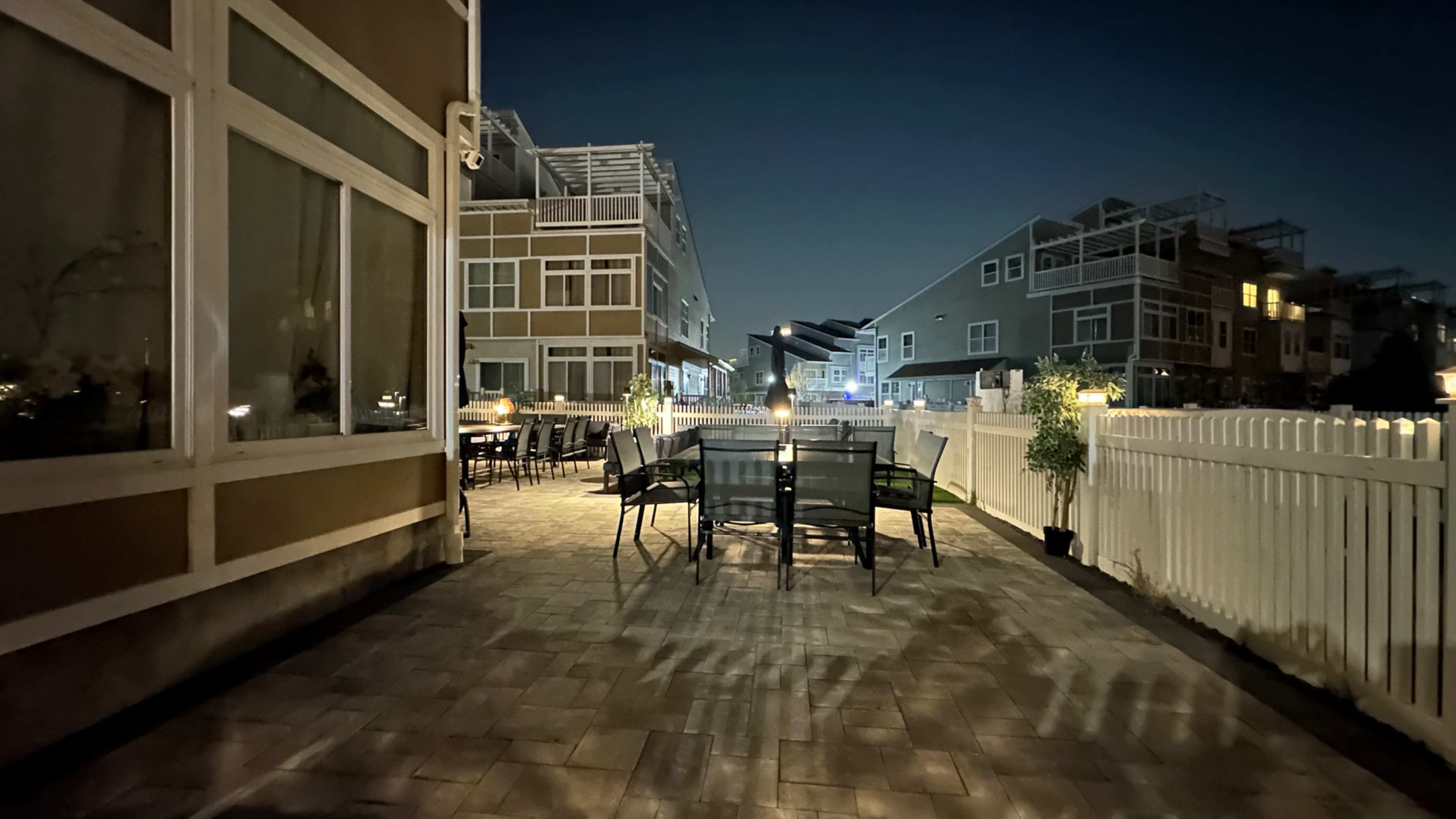A patio area surrounded by buildings is illuminated at night, featuring metal tables and chairs, a fence, and subtle shadows on the ground.