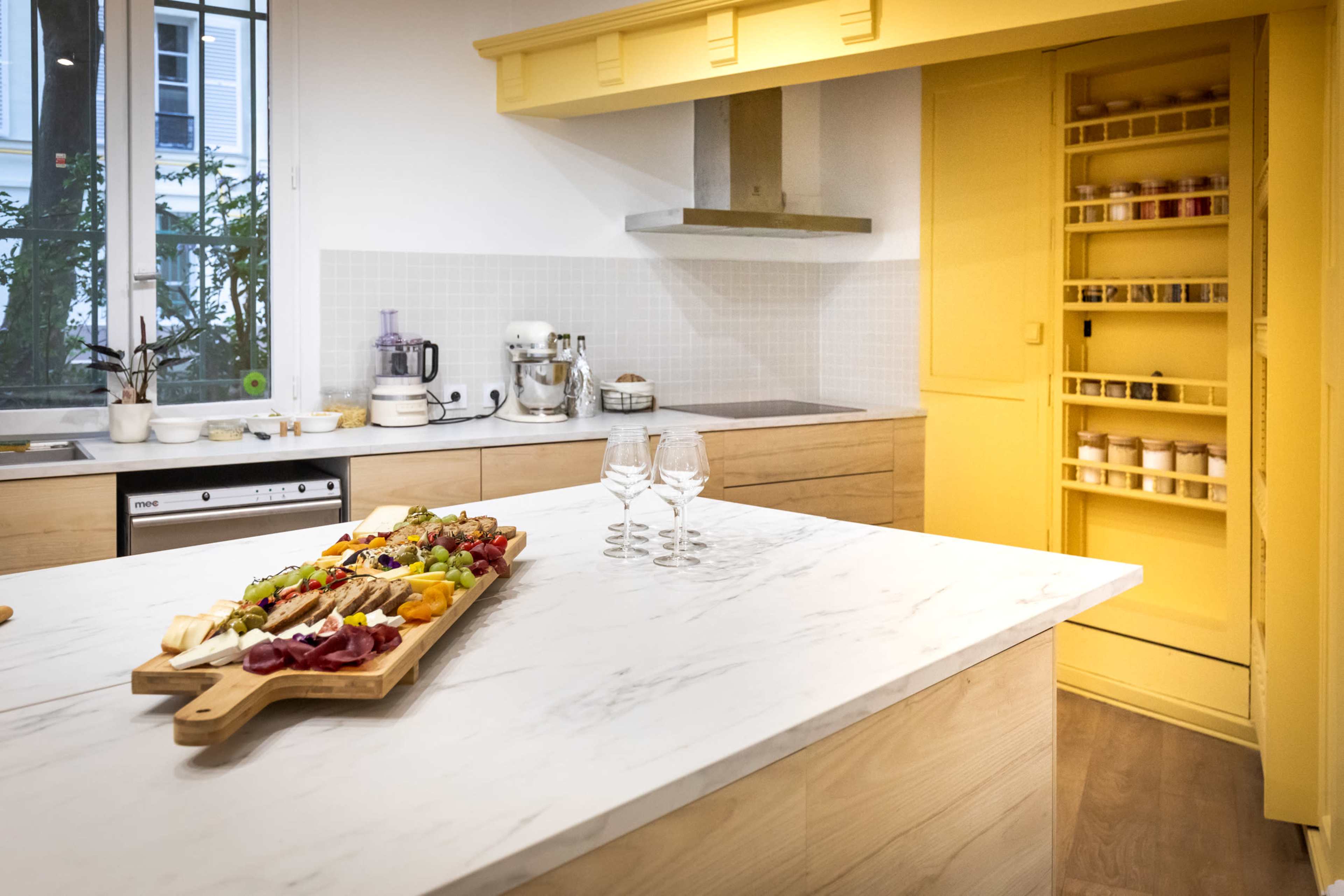 A bright kitchen features a marble island topped with a colorful charcuterie board, adjacent to a yellow cabinet and modern appliances.