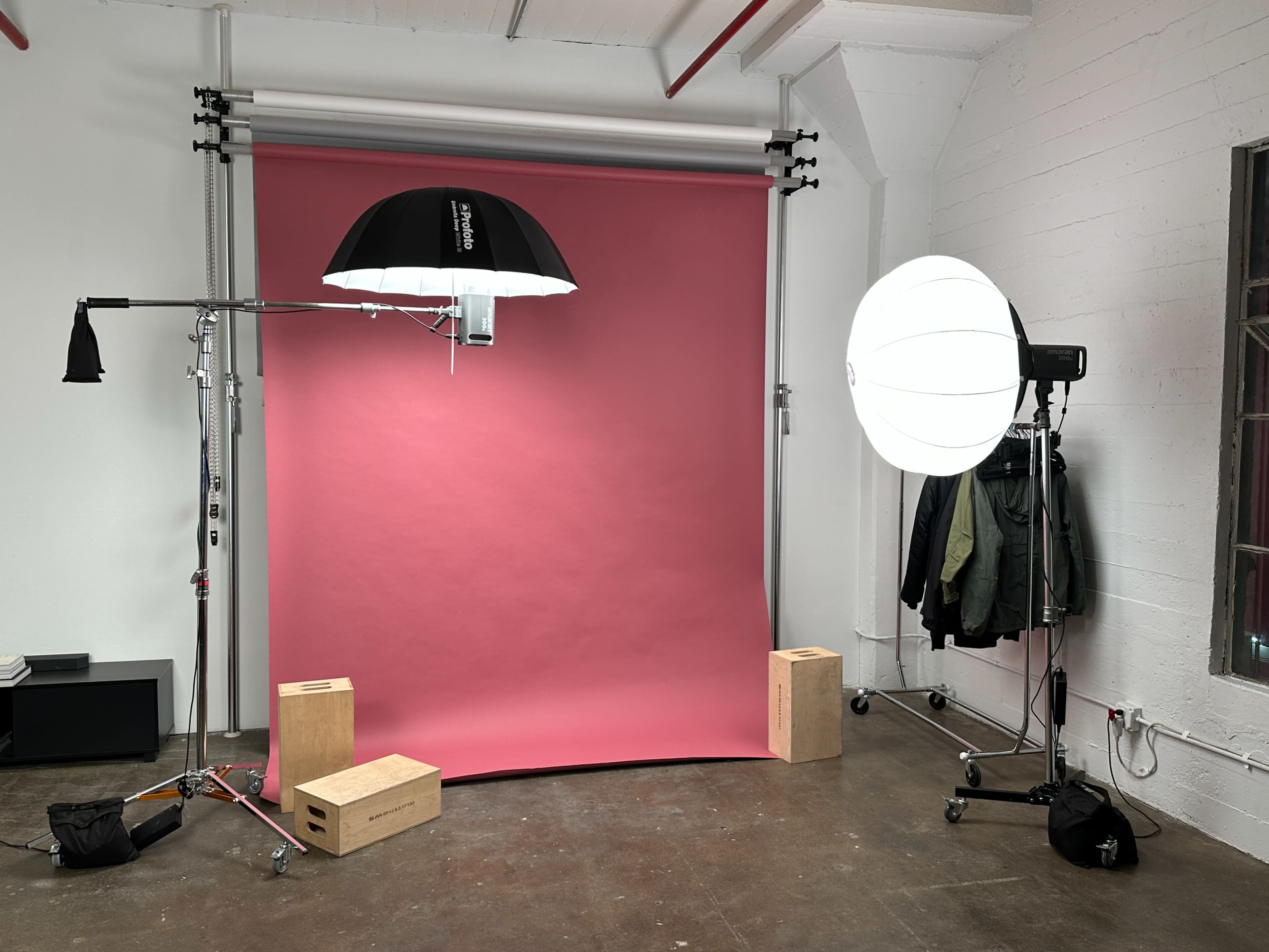 The image shows a photography studio setup with a pink backdrop, two lighting equipment stands, and wooden boxes on the floor.