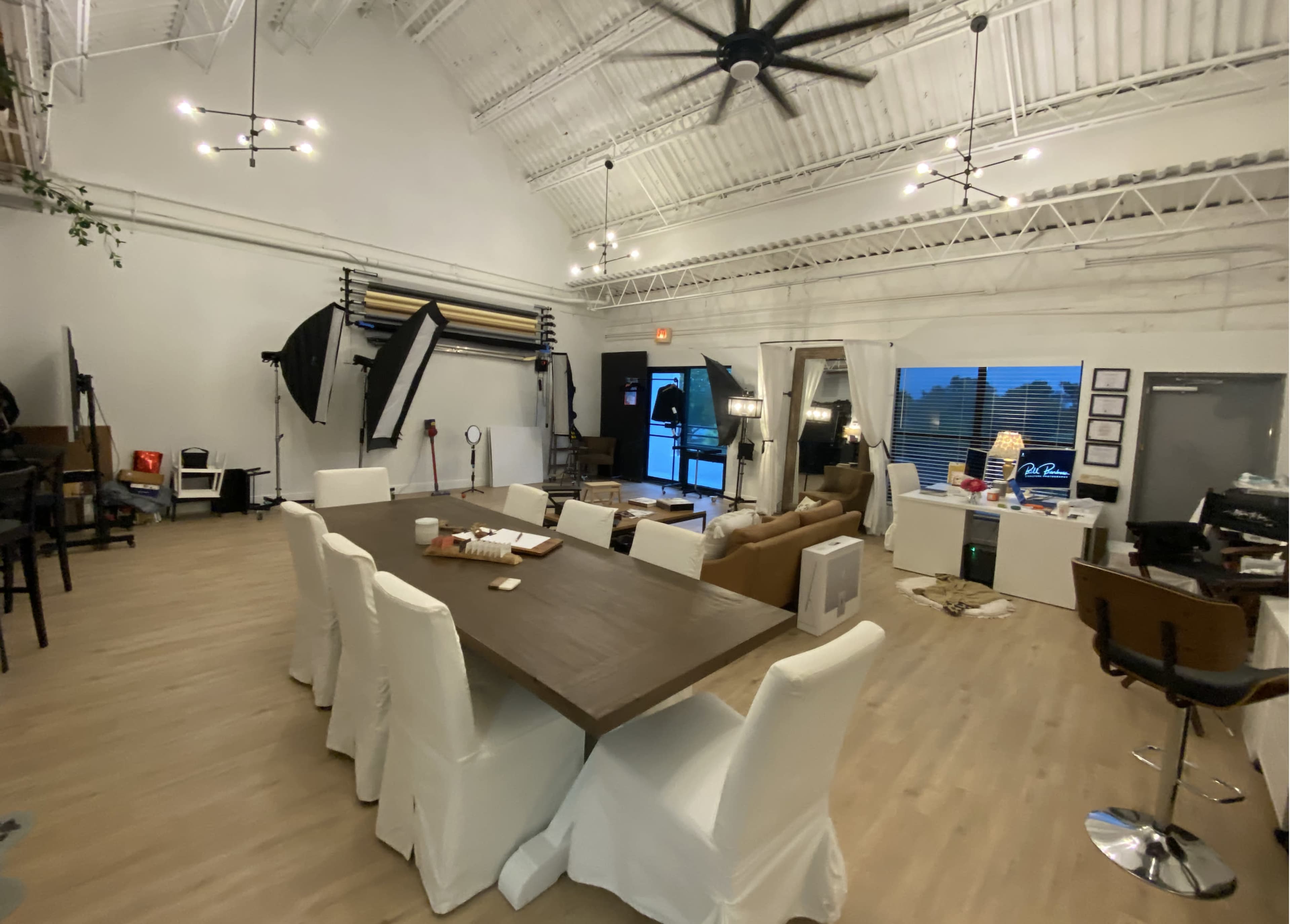 The image shows a spacious studio interior with a large table surrounded by white chairs, various photography equipment in the background, and large windows letting in natural light.