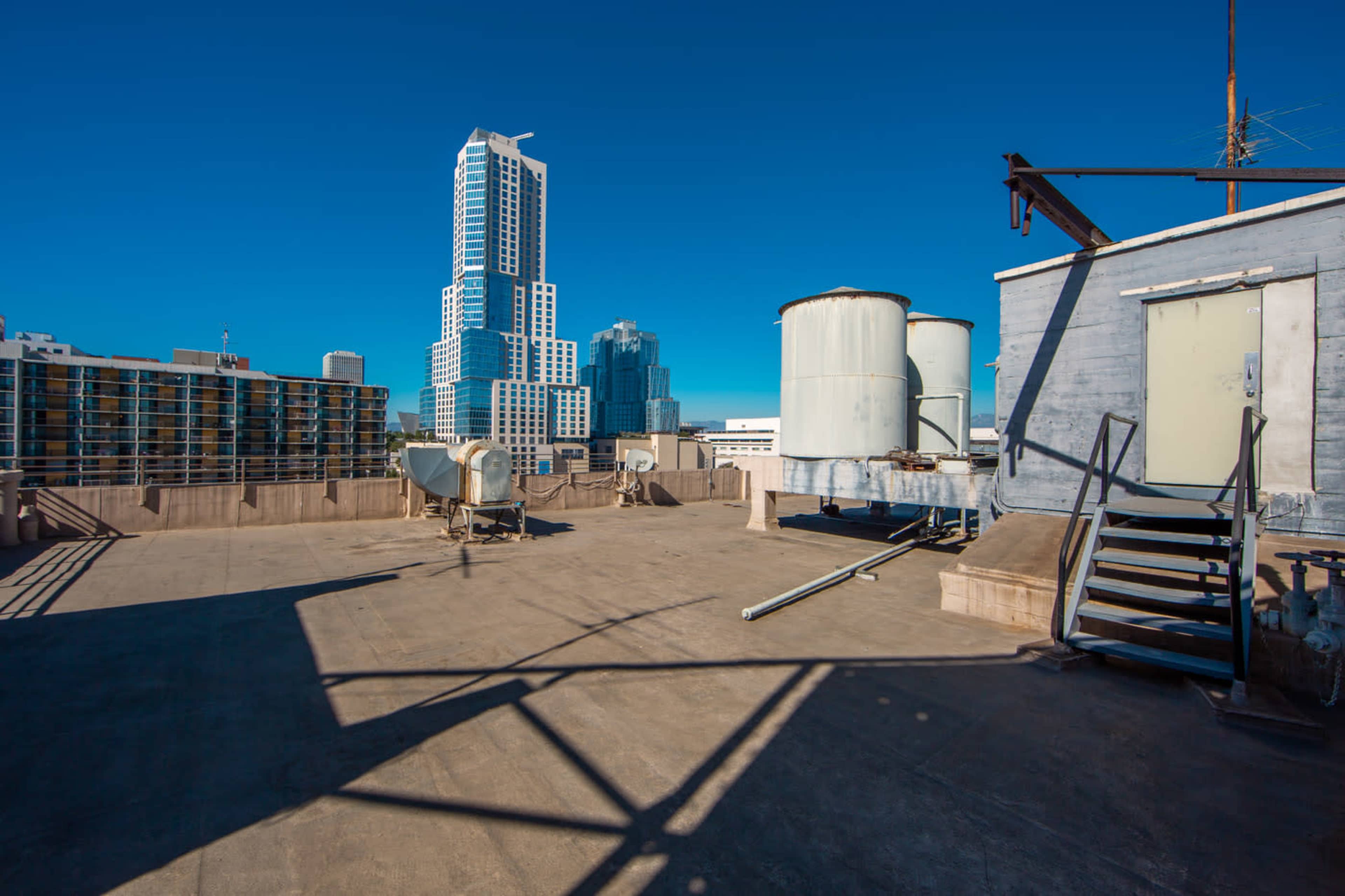 The image shows a rooftop view featuring large water tanks and a metal staircase, with a modern high-rise building in the background against a clear blue sky.
