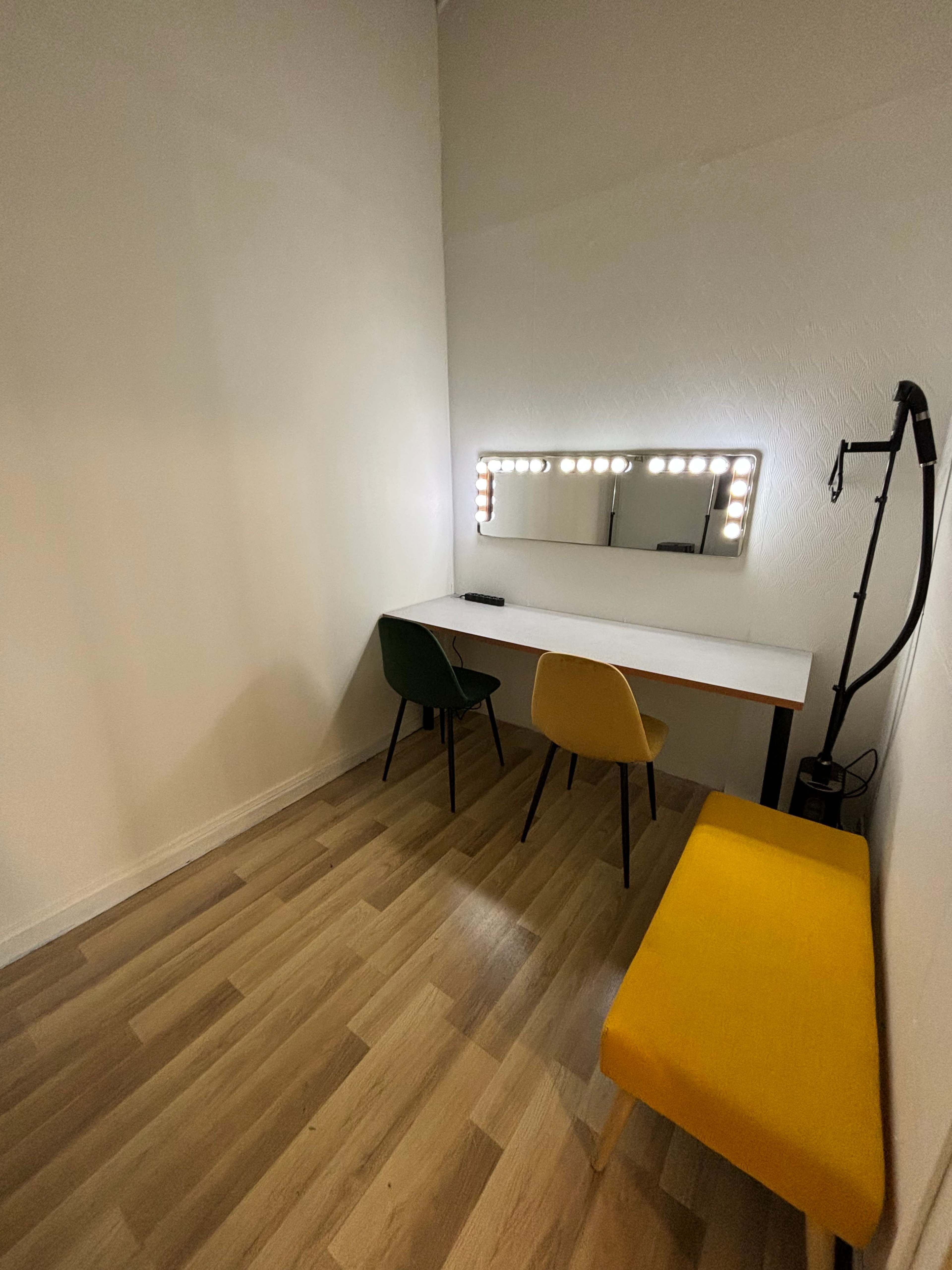 A small room with a table, a mirror surrounded by lights, two chairs, and a yellow bench along the wall.