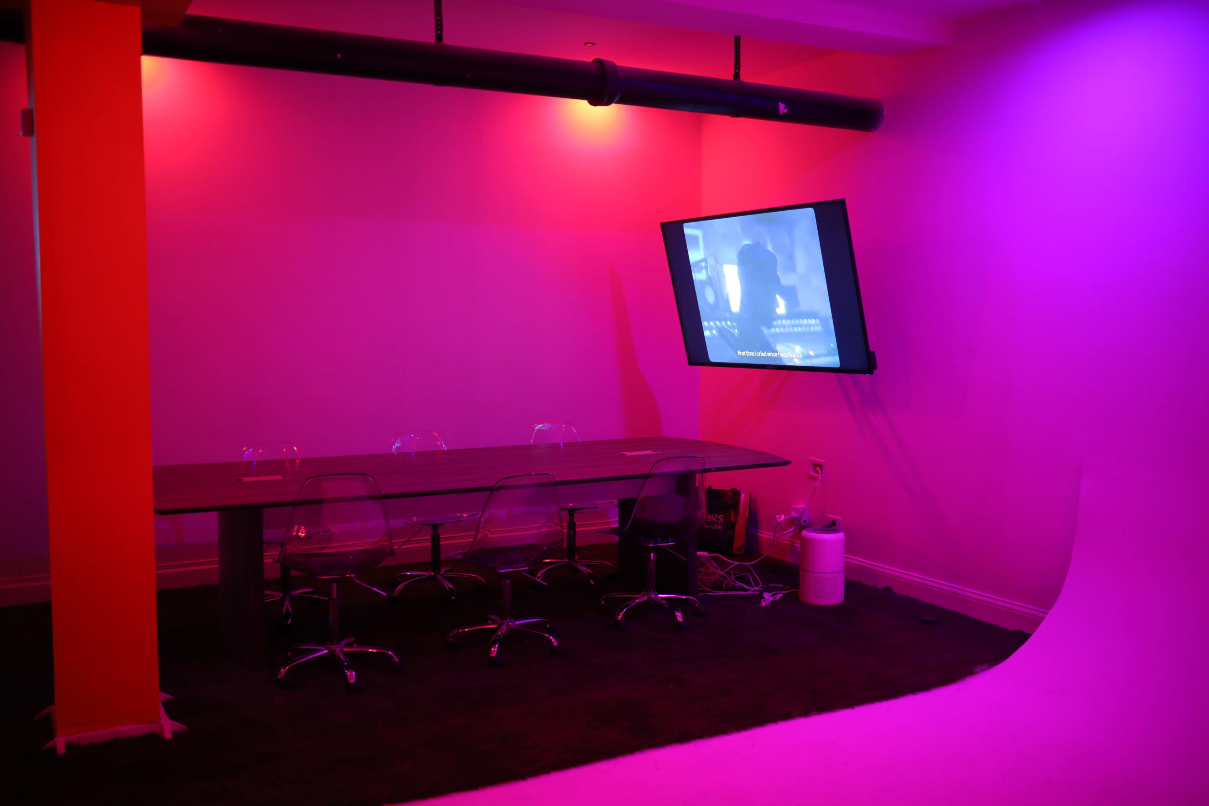 A dimly lit room features a large rectangular table surrounded by clear chairs, with a wall-mounted screen displaying a projection.