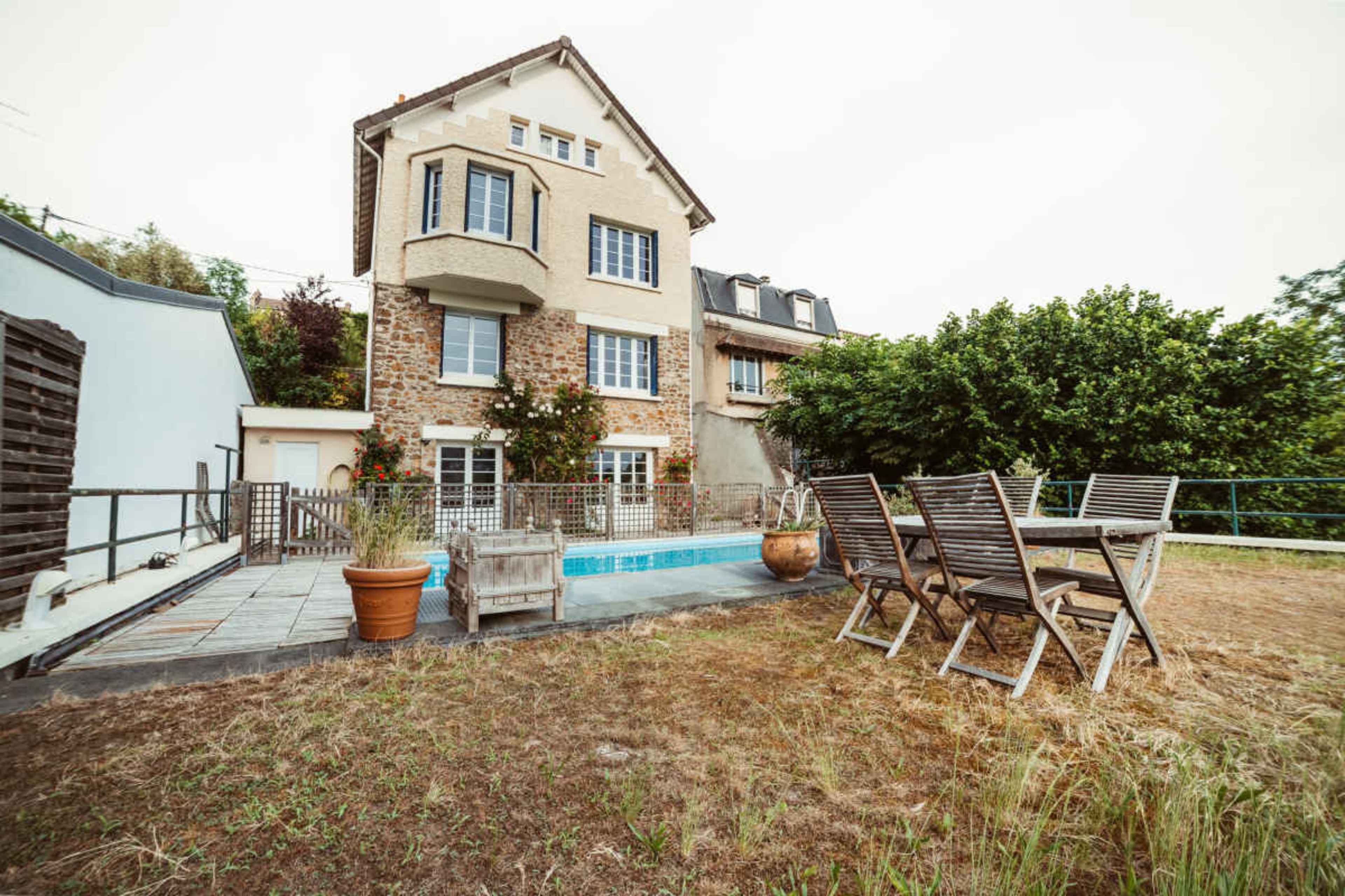 The image shows a three-story house with a stone facade, a swimming pool, and a wooden dining set in a grassy yard.