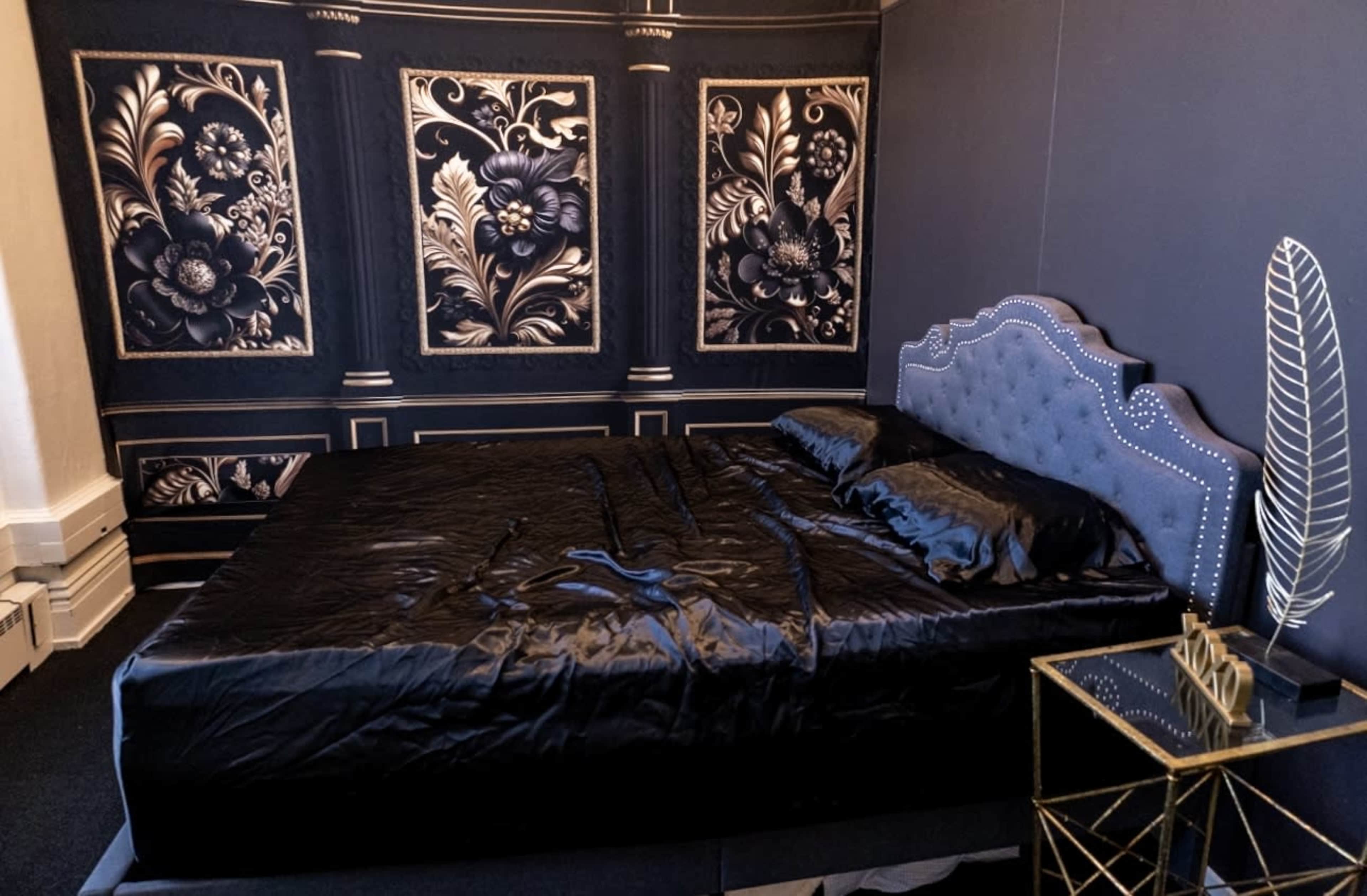 The image shows a bedroom with a black bedspread on a bed, against a wall featuring ornate floral patterns in black and gold.