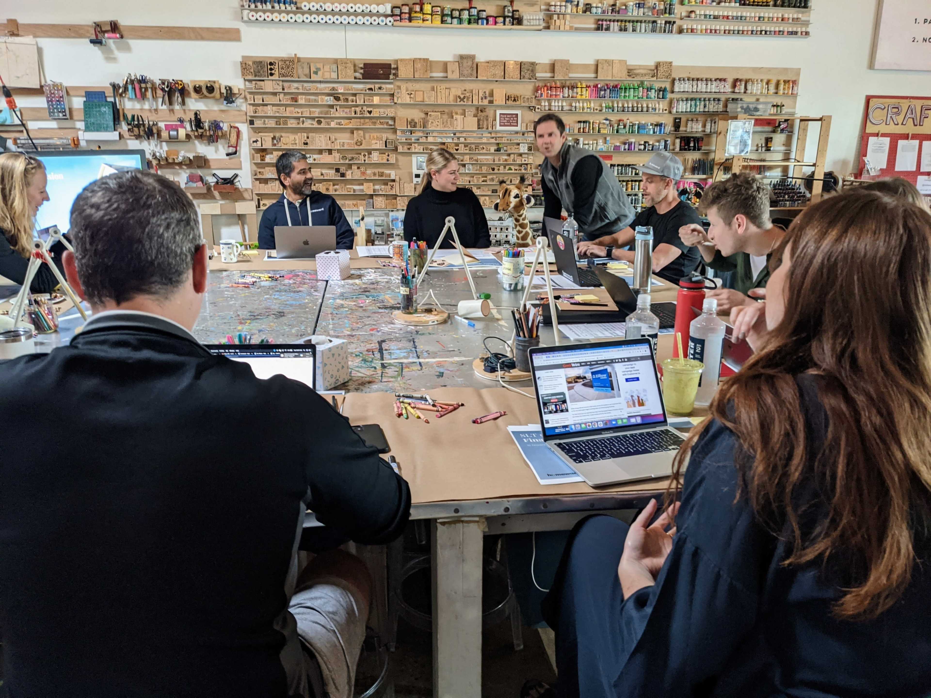 A group of people is gathered around a large table filled with laptops and craft supplies, engaged in discussion in a workshop setting.