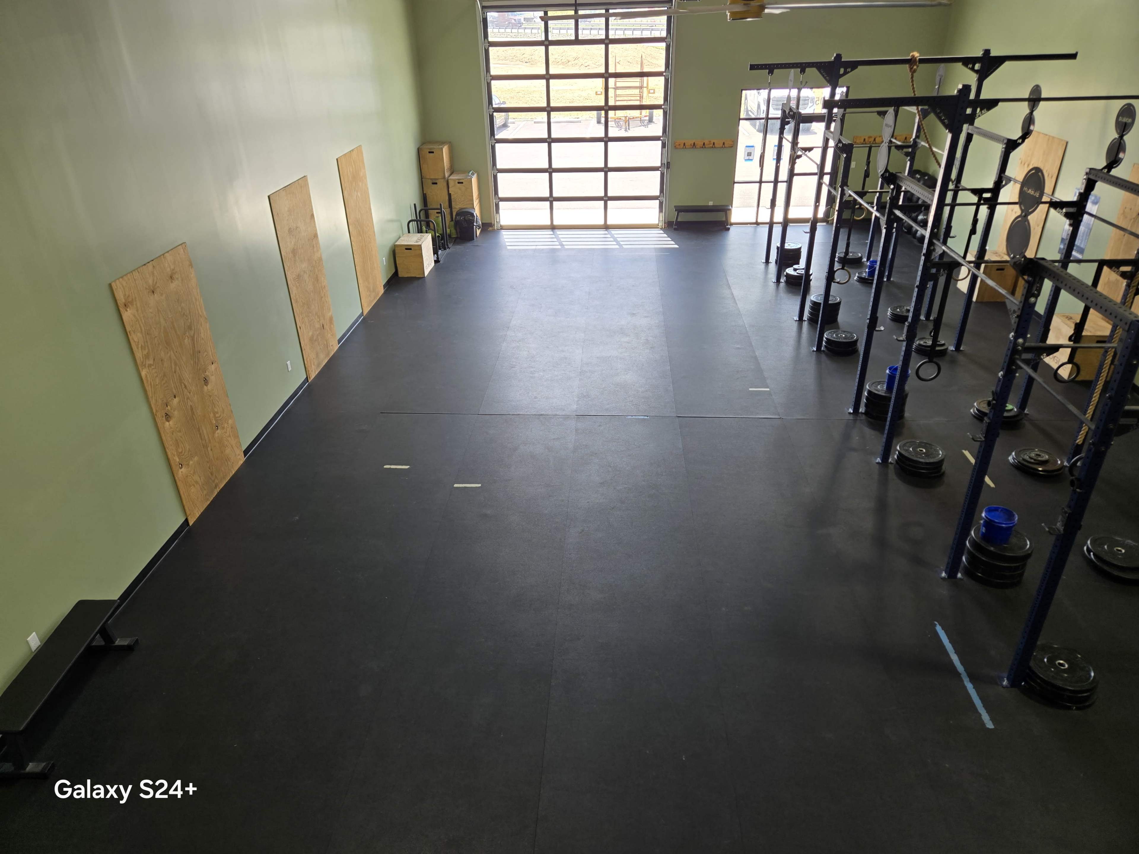 The image shows an empty gym space with black flooring, wooden panels on the walls, and weightlifting equipment arranged in an orderly manner near large windows.