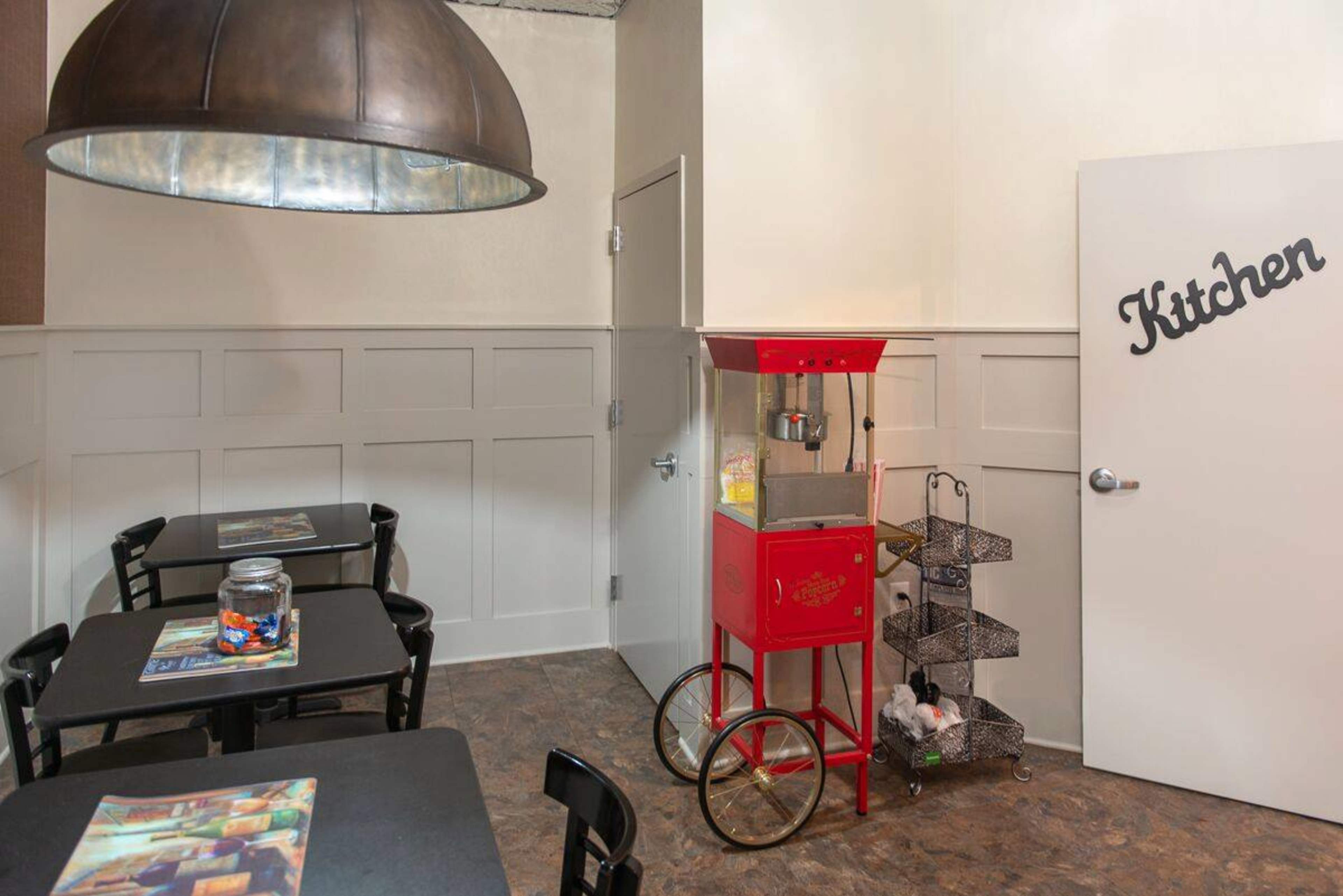 The image shows a corner of a room featuring a red popcorn machine on a wheeled cart, a large pendant light hanging above, and a door marked "Kitchen."