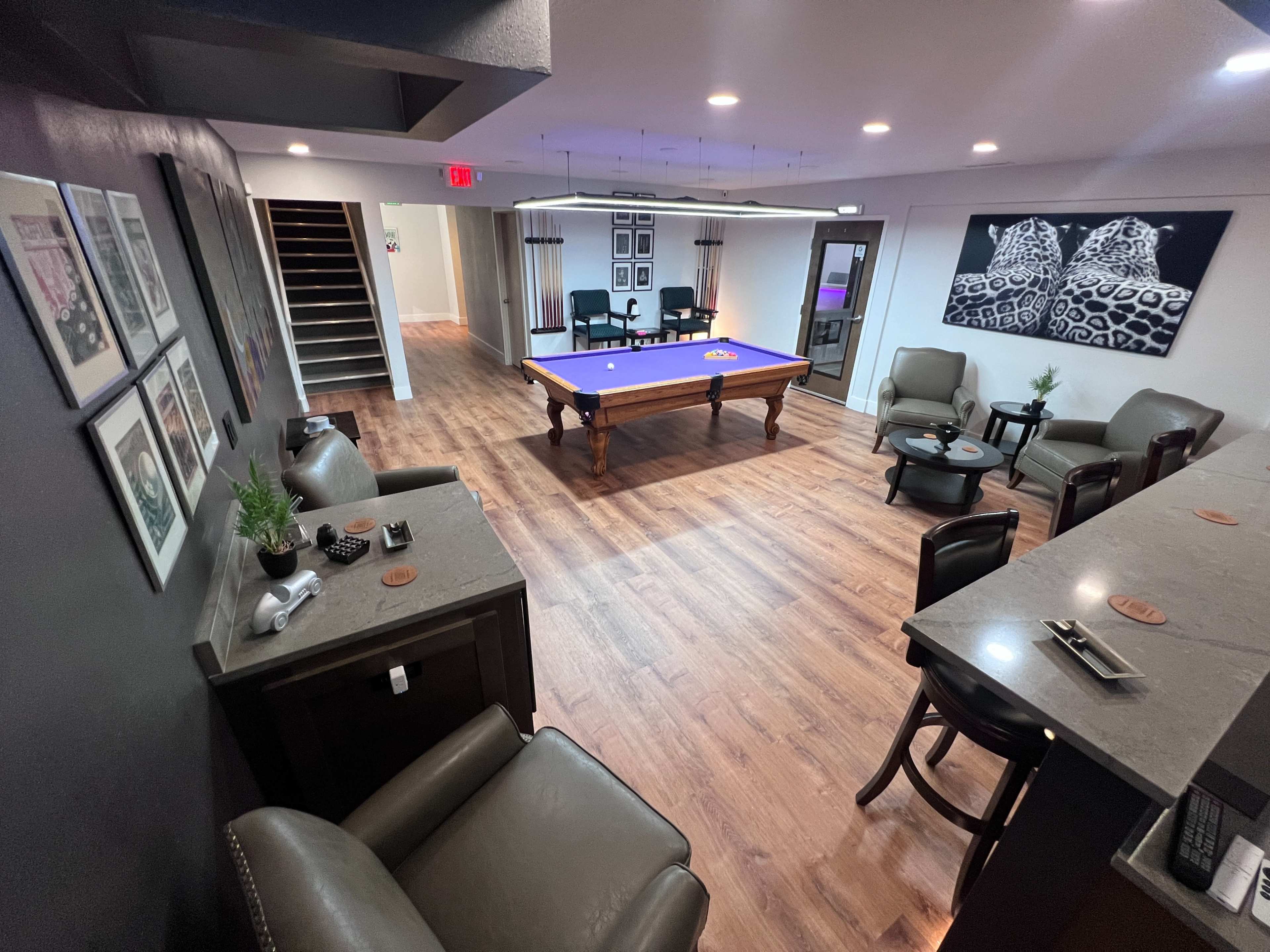 A modern recreational room featuring a pool table at its center, surrounded by seating areas and a bar.
