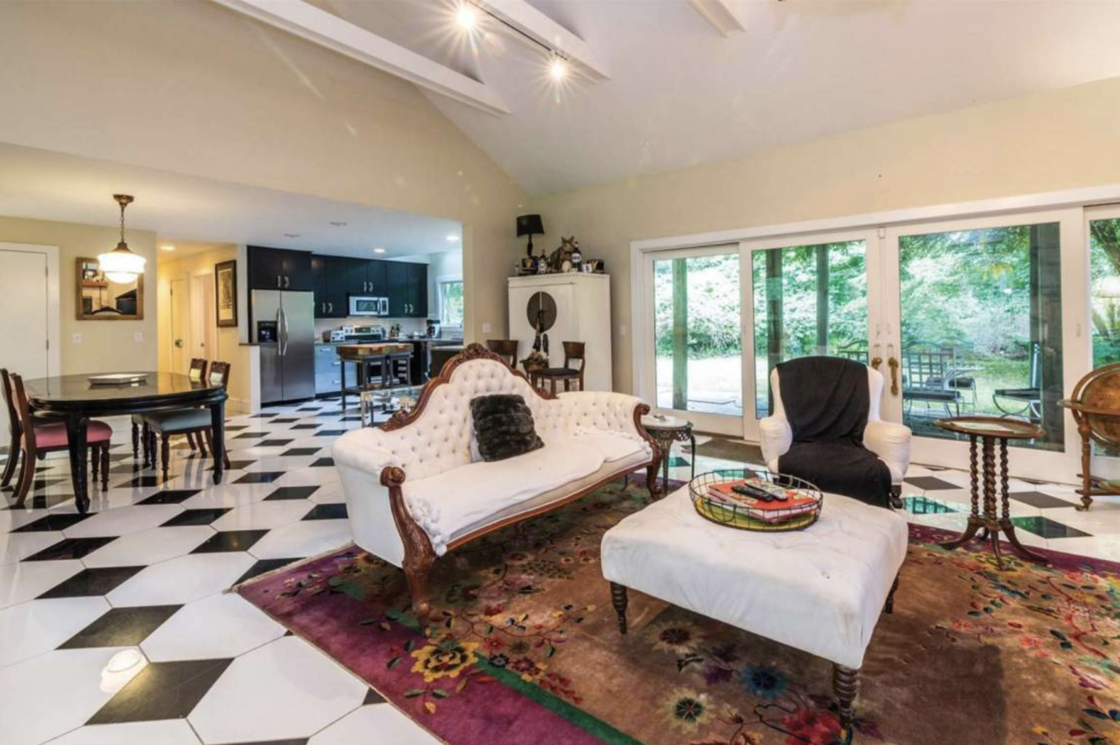 A spacious living room with a checkered black-and-white floor, a white tufted couch, and a large window overlooking a garden.