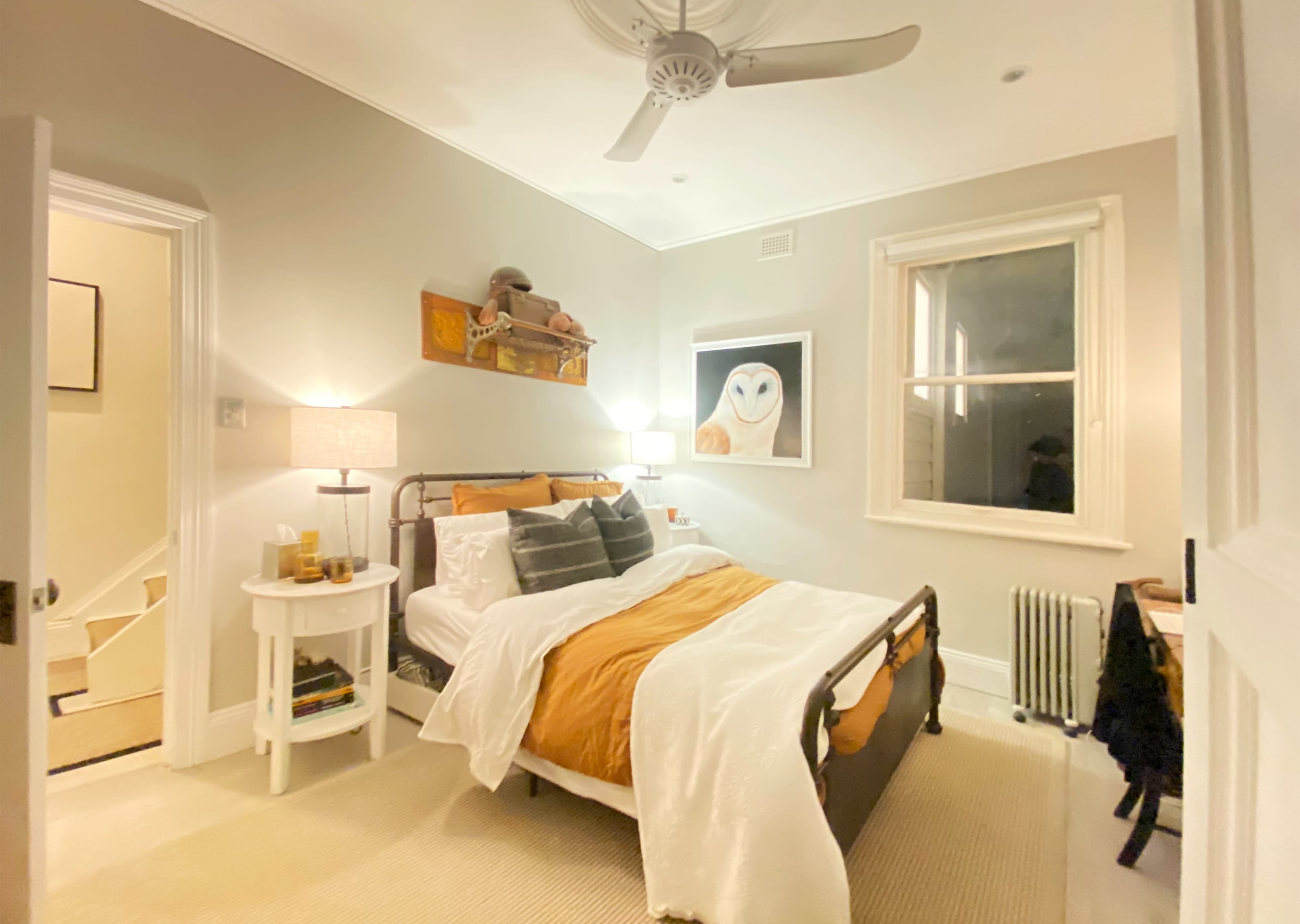 A neatly arranged bedroom features a bed with orange and white bedding, a bedside table, a window with drapes, and artwork of an owl on the wall.