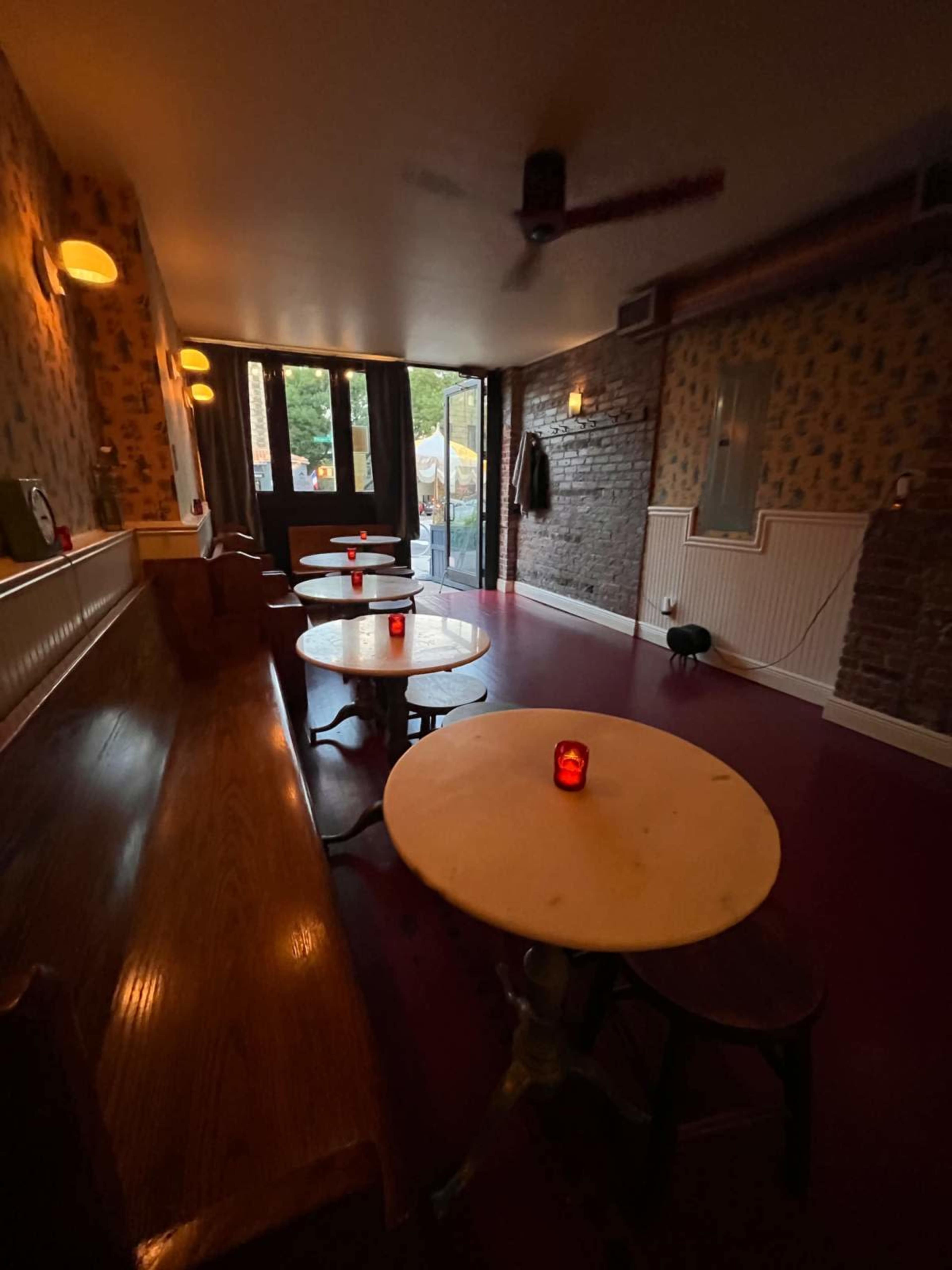 The image shows a dimly lit room with wooden tables and benches, featuring red candles on each table and a view of an outdoor area through large windows.