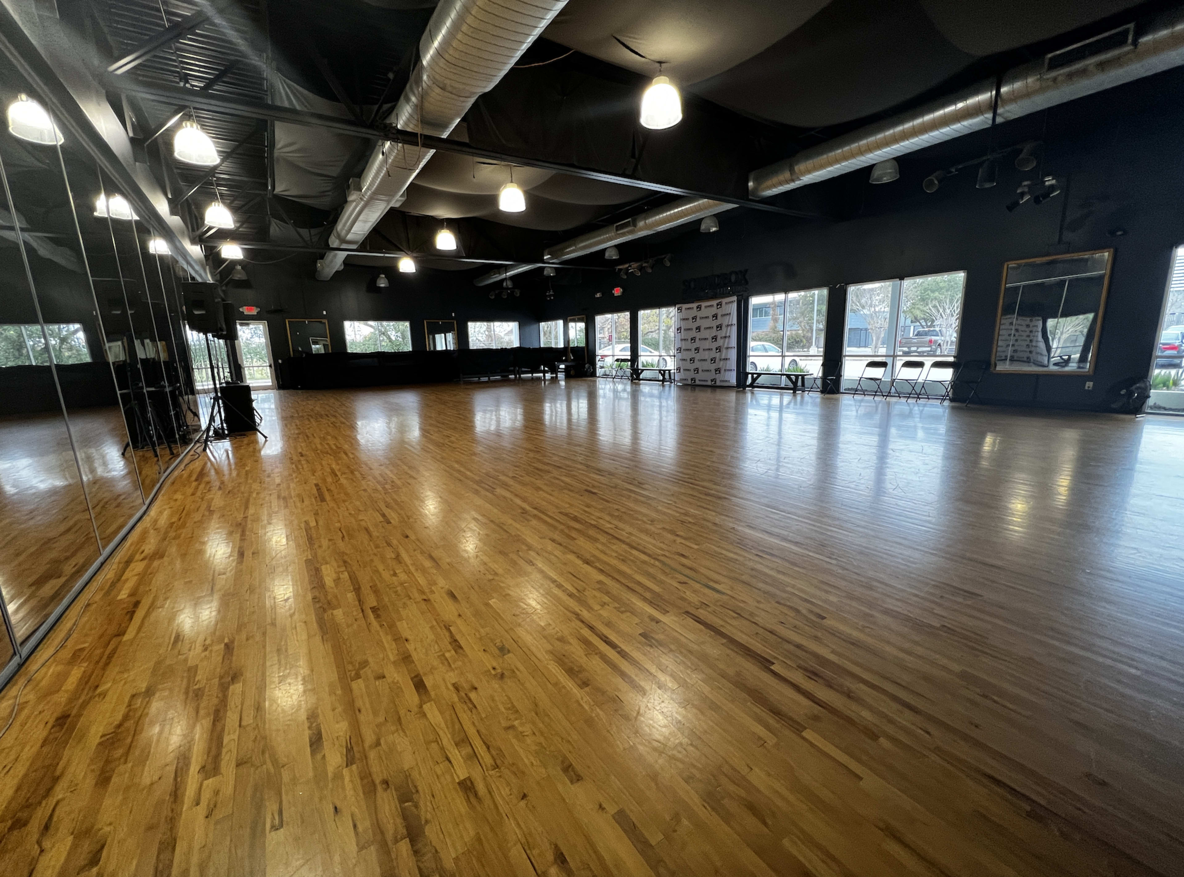 A spacious dance studio with a polished wooden floor, large mirrors, and windows that allow natural light to enter.