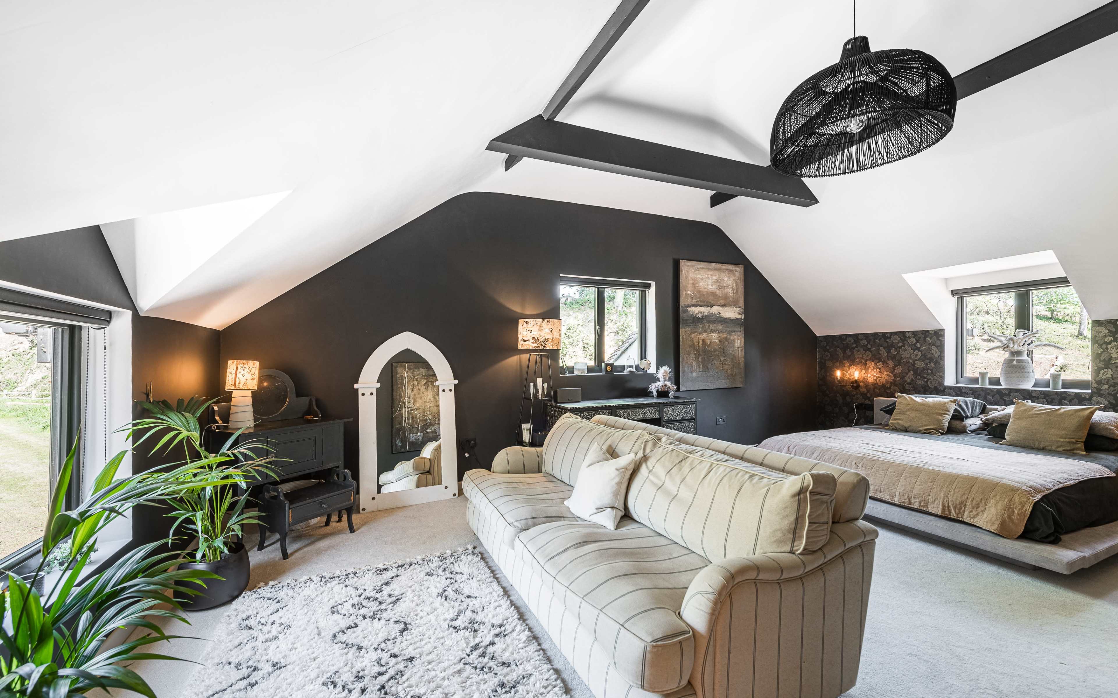 A spacious bedroom features a large bed, a sofa, a decorative mirror, and large windows that provide natural light.