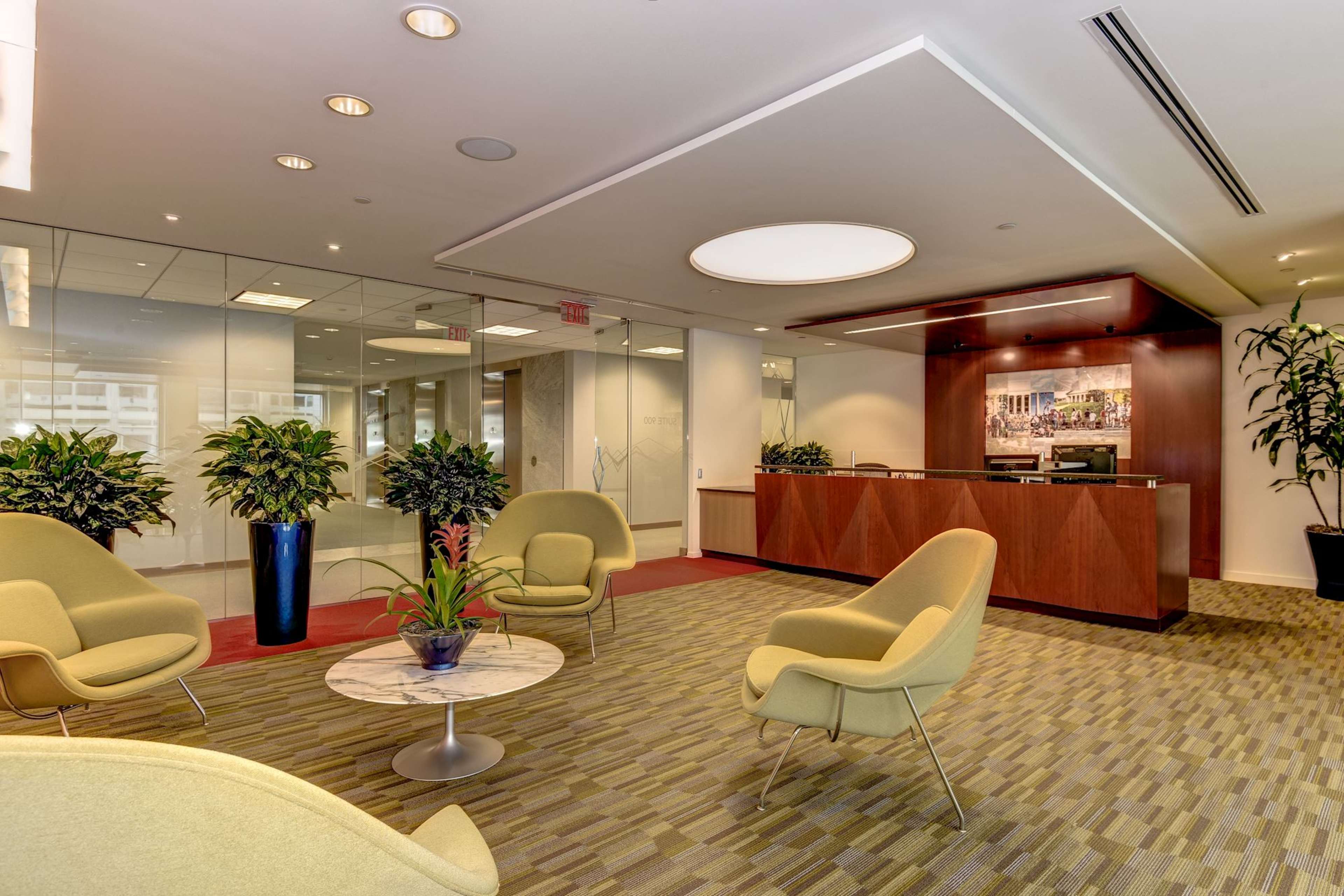 The image shows a modern reception area with a marble table, green chairs, and a wooden front desk, surrounded by potted plants.