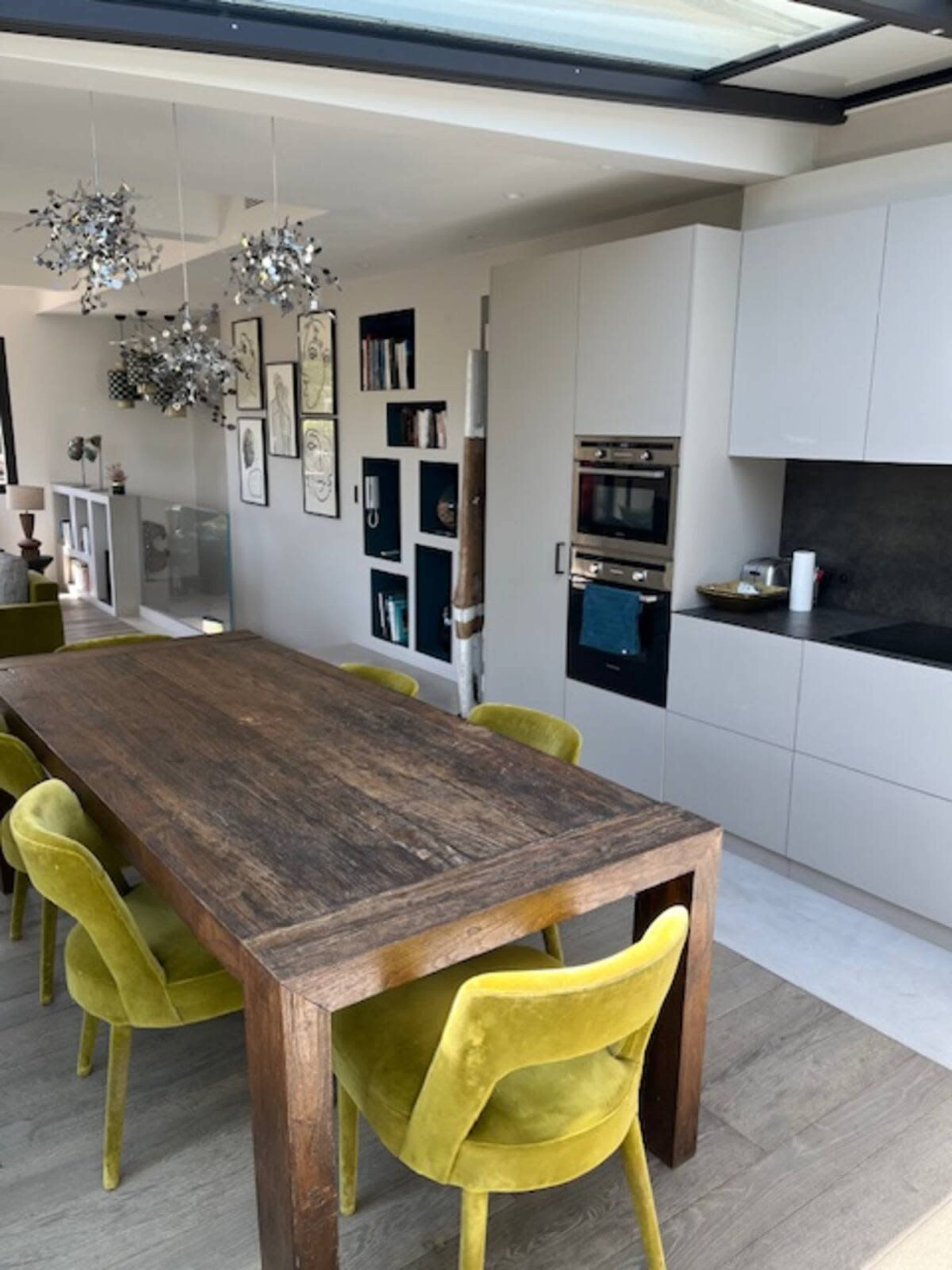 The image shows a modern dining area with a large wooden table surrounded by several yellow chairs, and a sleek kitchen with integrated appliances in the background.