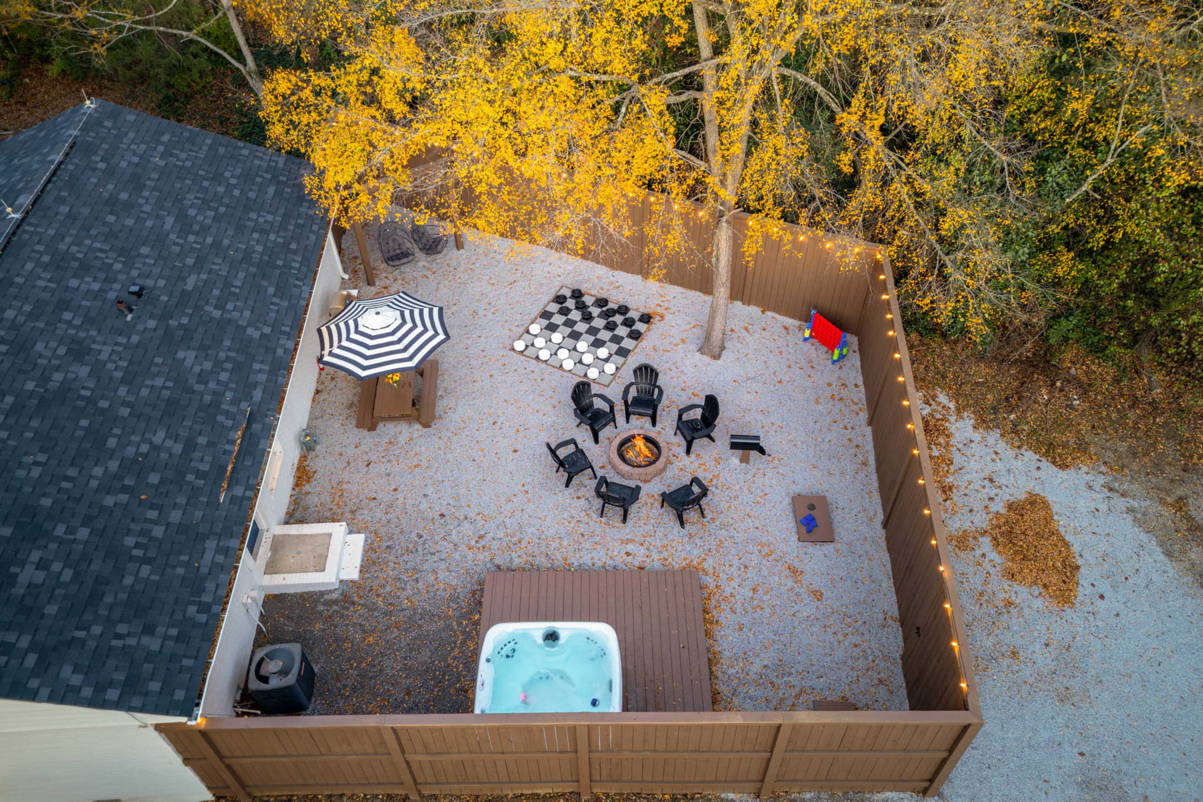 The image shows an aerial view of a backyard featuring a hot tub, a fire pit surrounded by black chairs, a checkerboard area, and a large umbrella, all set within a fenced area with fall foliage.