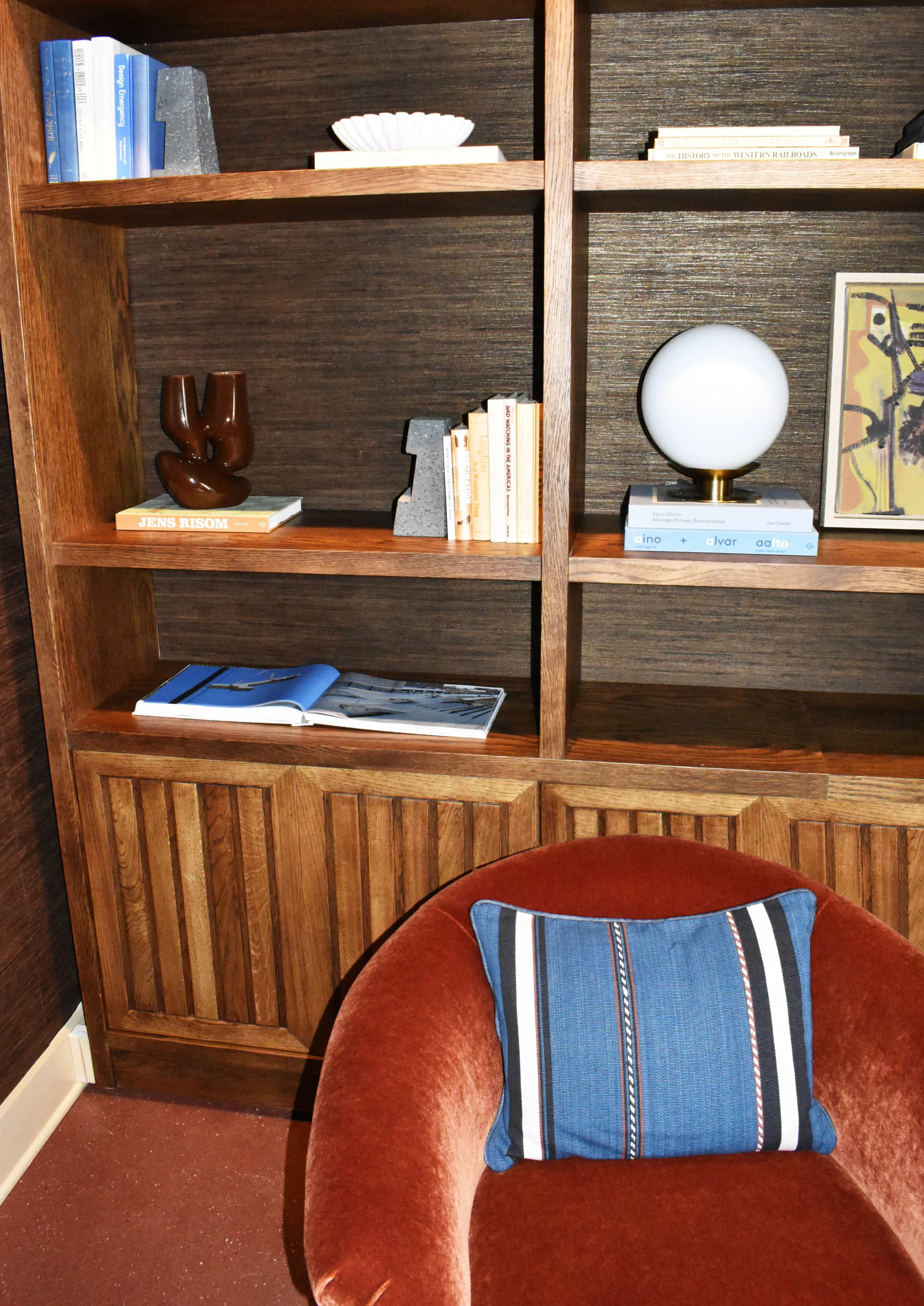 The image shows a wooden bookshelf filled with books, decorative items, and a round lamp, alongside a red armchair with a blue striped cushion.