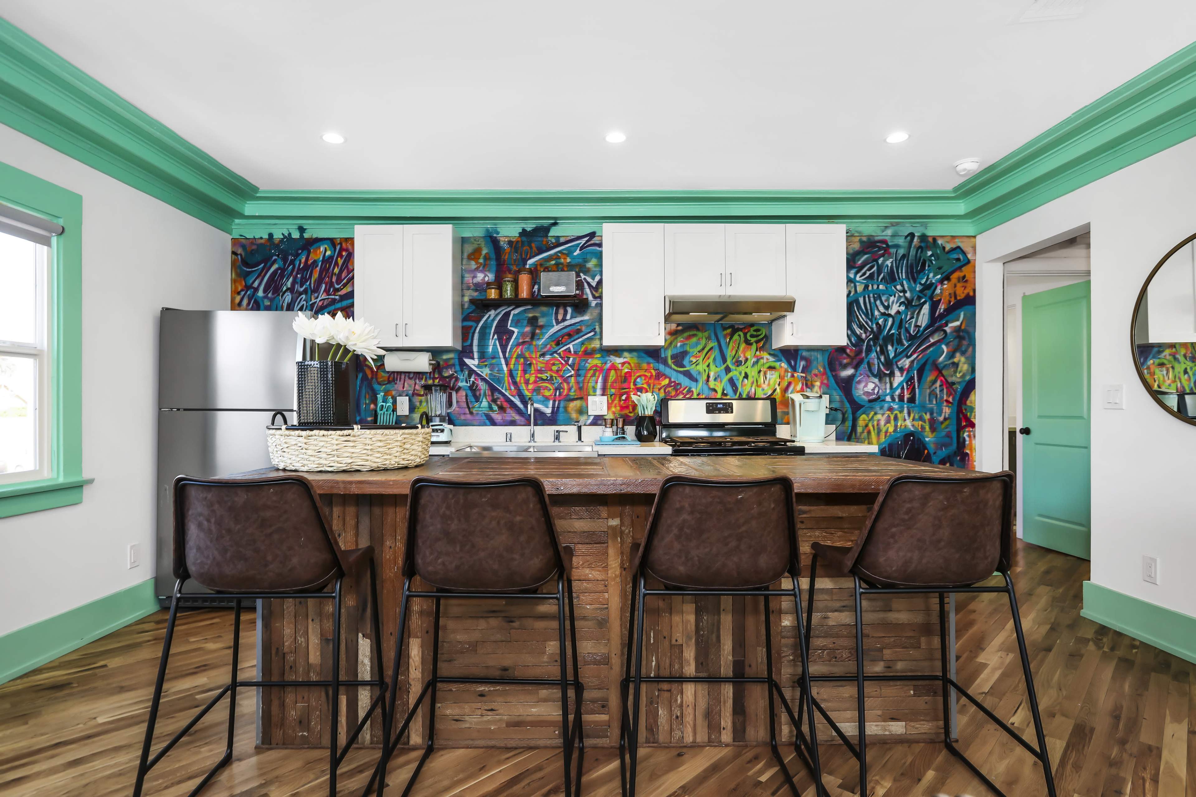 A modern kitchen features a wooden island with four high stools, white cabinetry, and a vibrant graffiti mural on the wall.