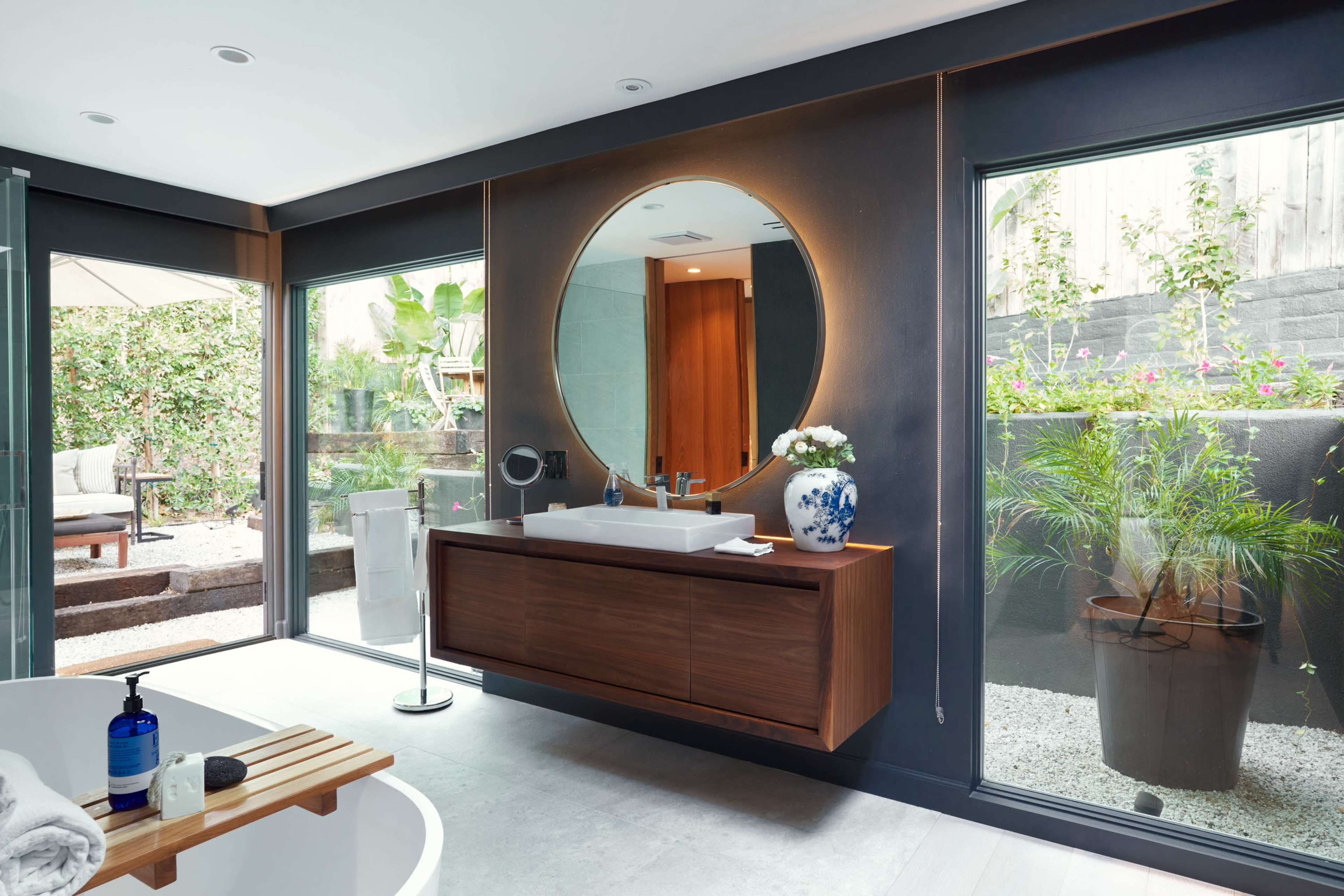 The bathroom features a round mirror above a wooden vanity, with large glass windows offering views of an outdoor garden area.