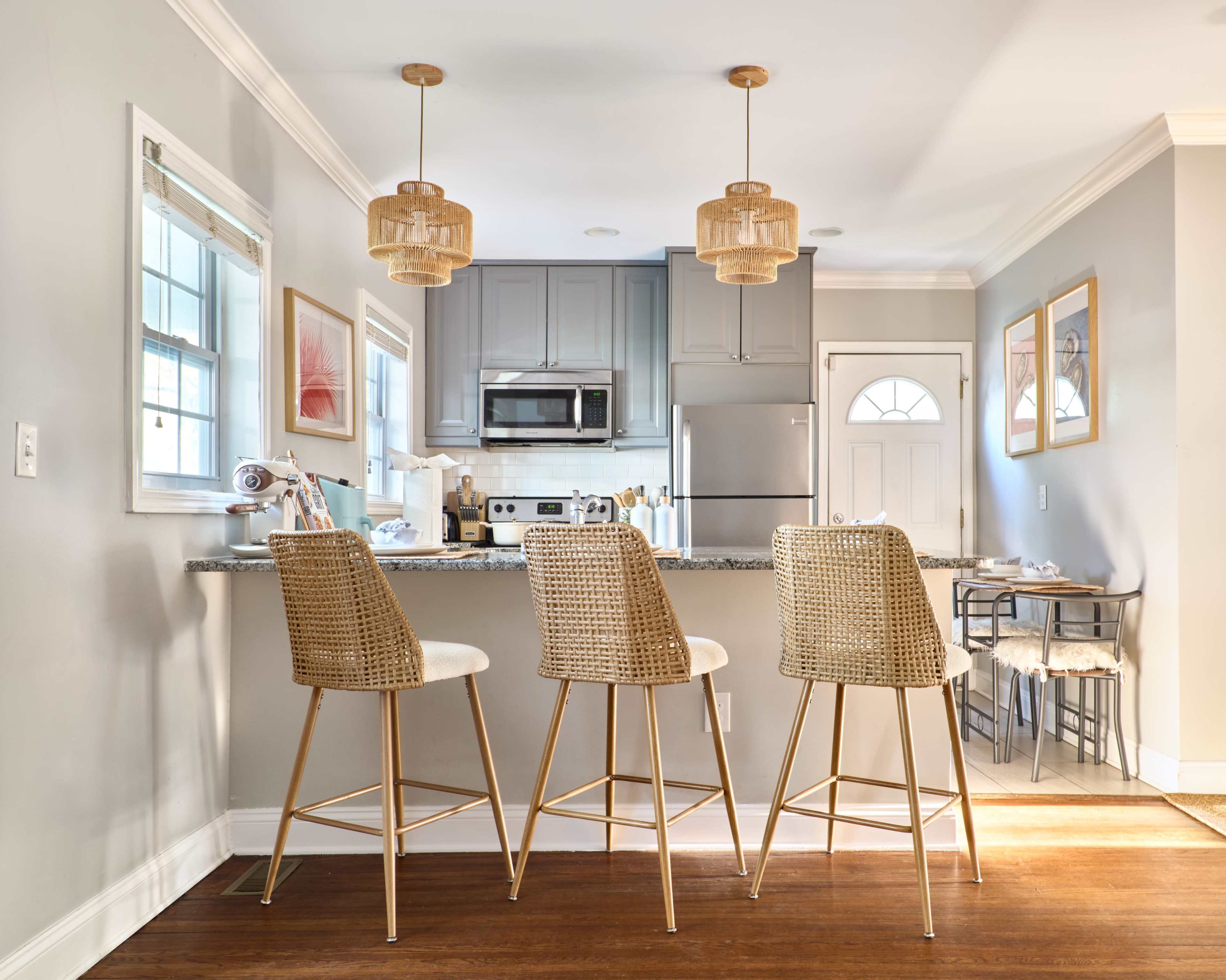 The image shows a bright and modern kitchen with three rattan bar stools lined up at a granite countertop, illuminated by pendant lights.