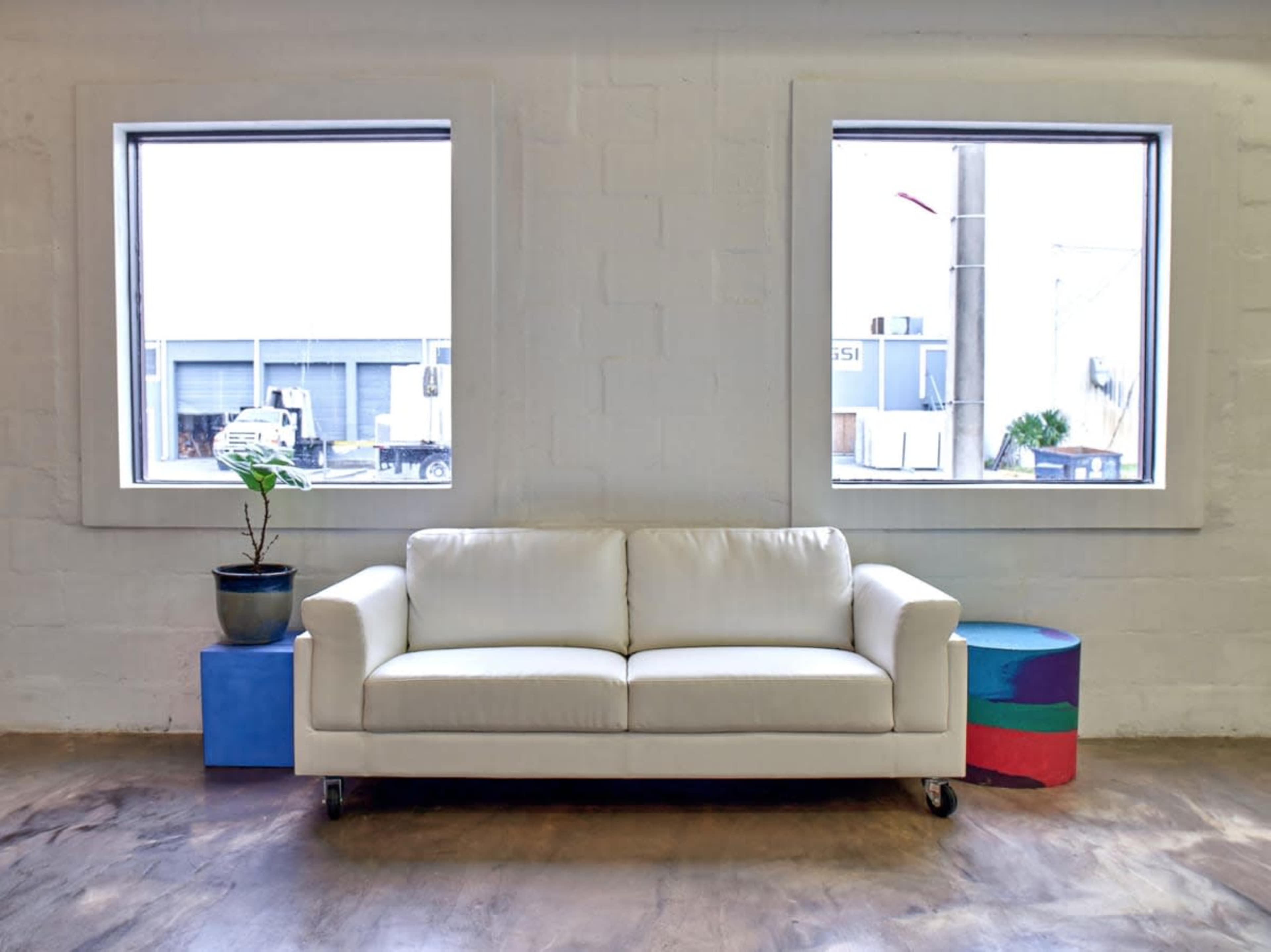 A white couch is positioned against a wall with two large windows, accompanied by a plant in a blue pot and a colorful cylindrical block.