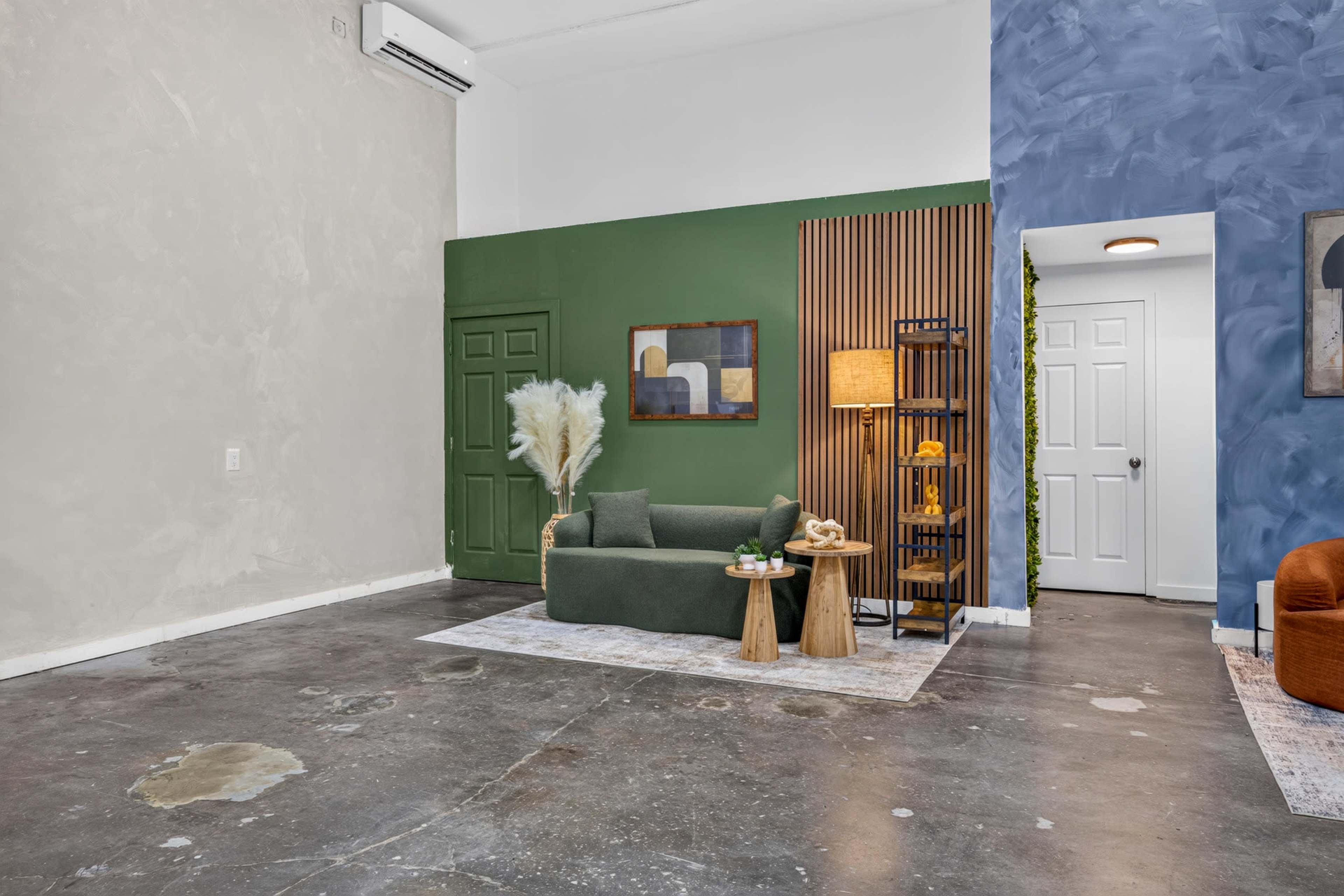 The image shows a spacious room with a green accent wall, a sofa, two wooden side tables, a lamp, and a textured blue wall in one corner.