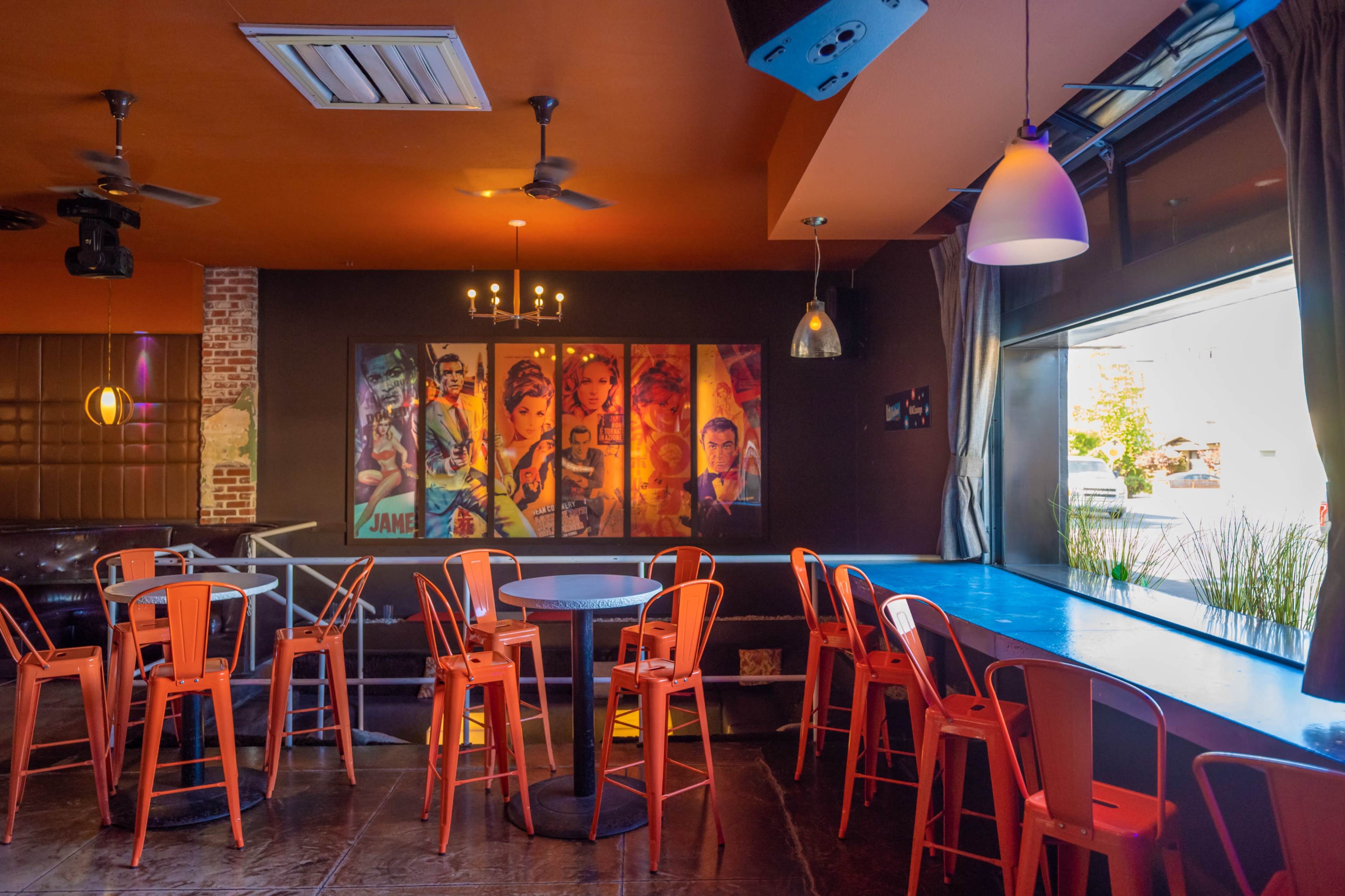 The image shows a modern cafe interior with orange metal chairs, a blue table, and colorful framed artwork on the wall.