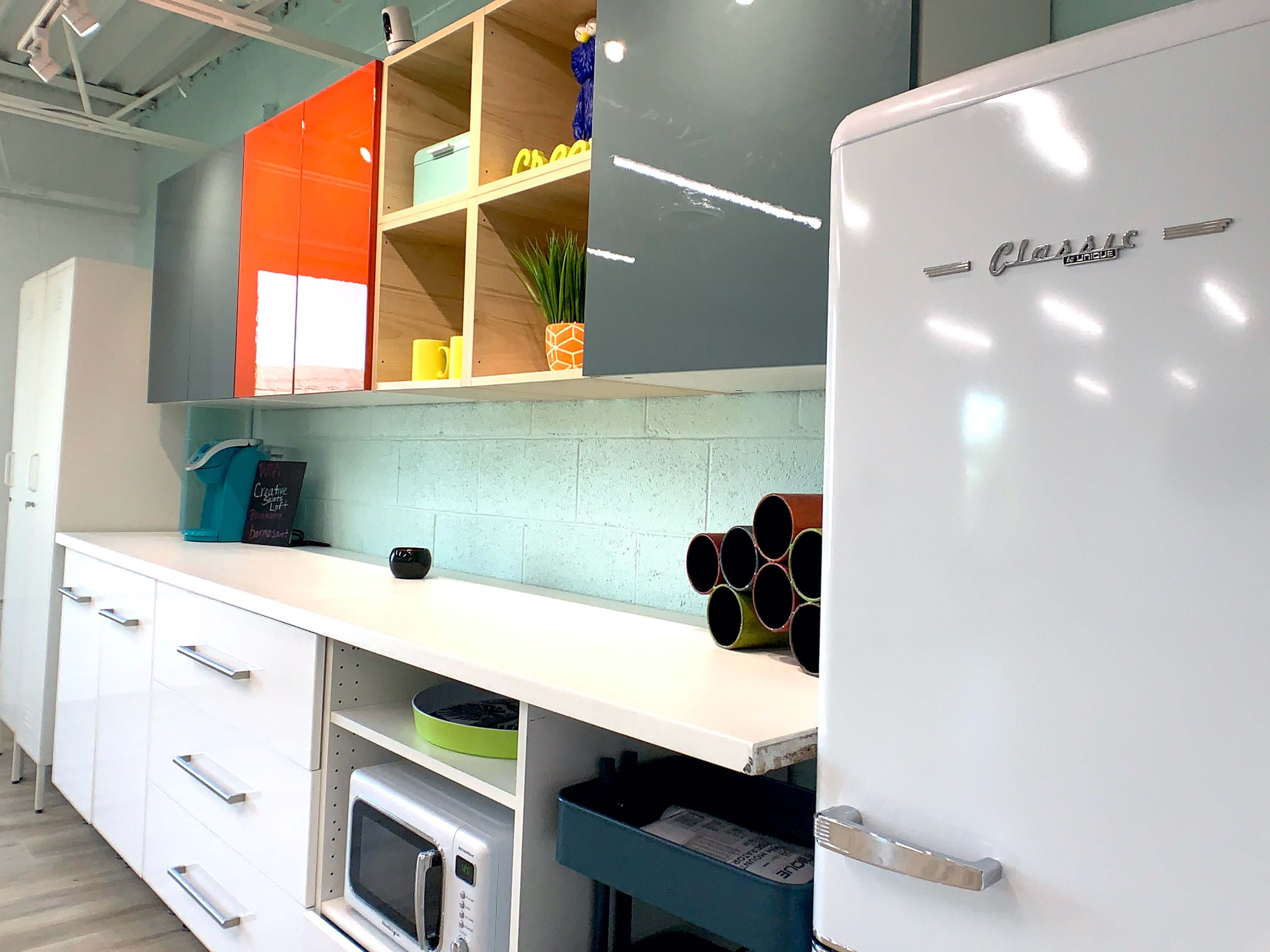 The image shows a modern kitchen interior with a white refrigerator, a microwave, and shelves displaying colorful items against a light-blue wall.