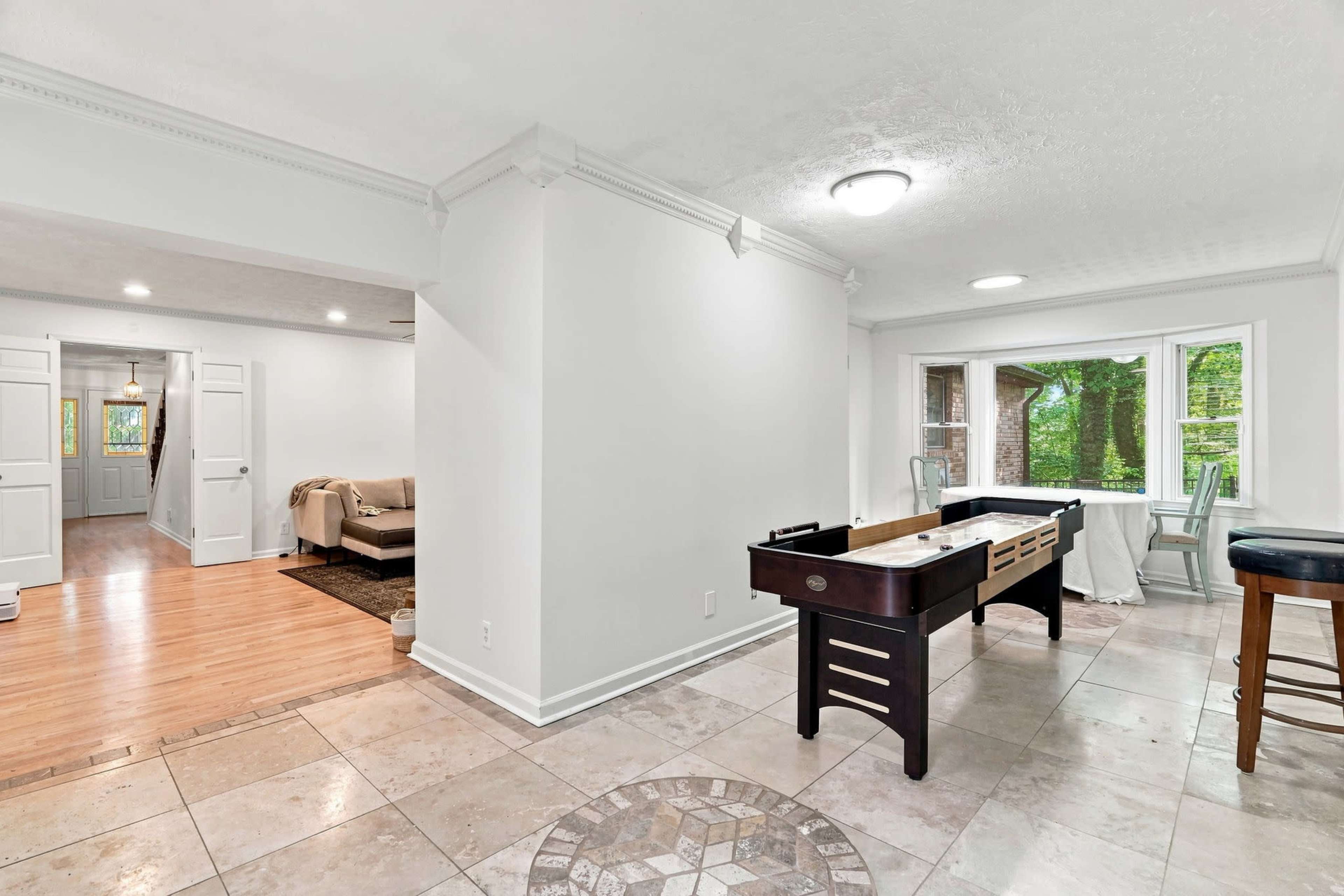 The image shows a spacious room with a pool table, tile flooring, and a view of a nearby window leading outside, alongside a doorway to another room.