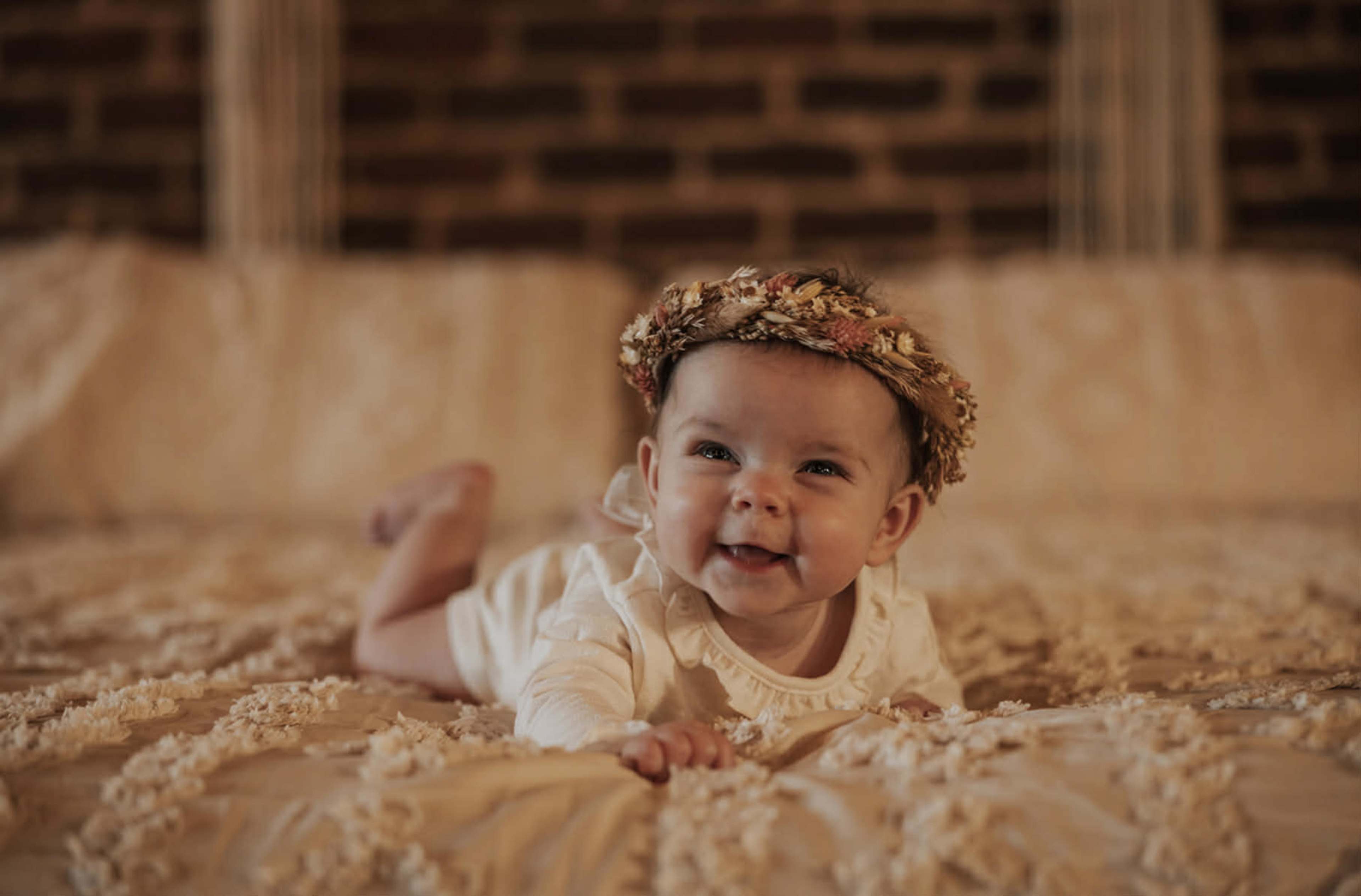 A baby wearing a floral headband is lying on a textured bedspread in a room with a brick wall.