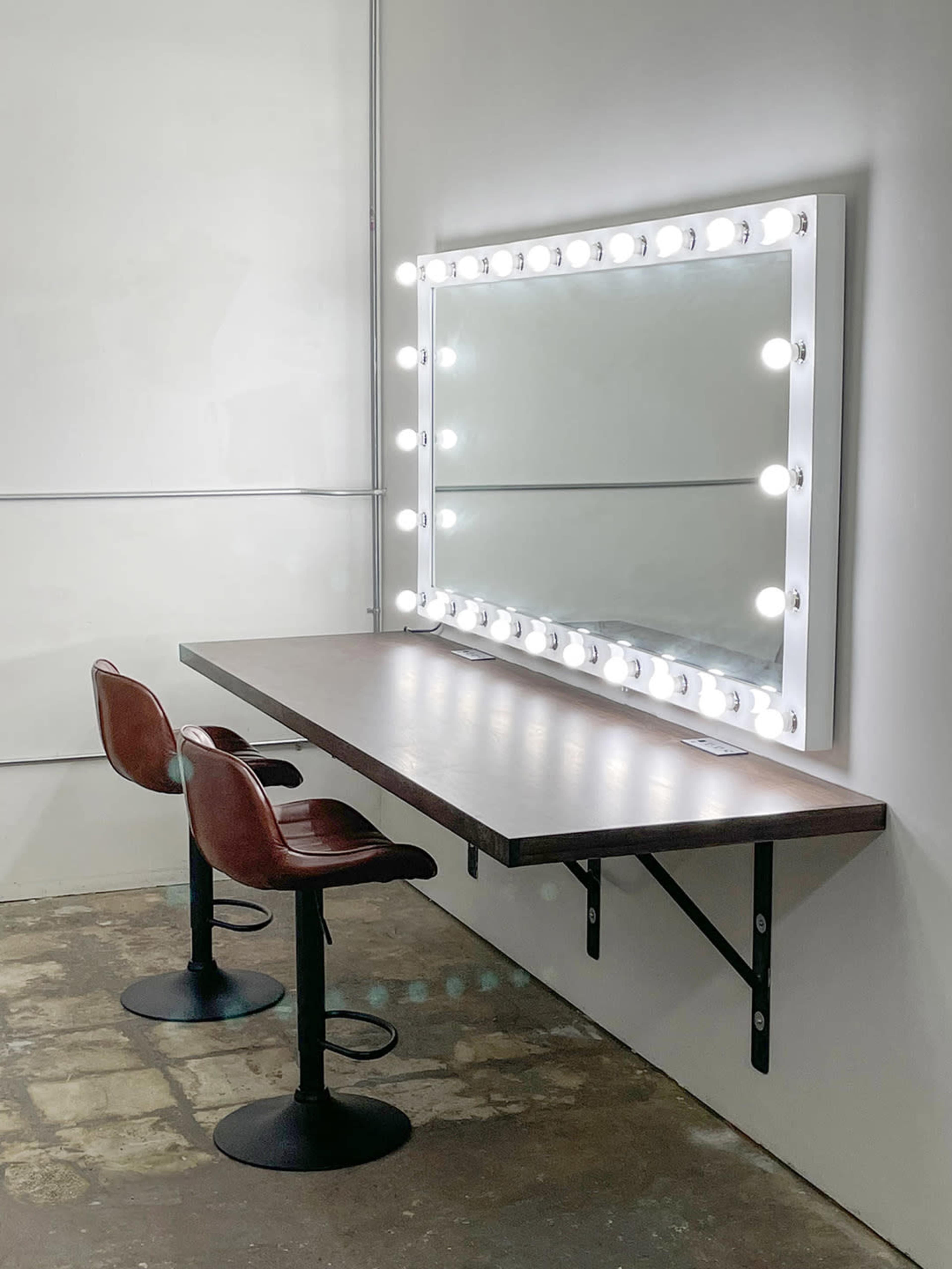 The image shows a minimalist makeup station with a large mirror surrounded by lights, a wooden table, and two red chairs.