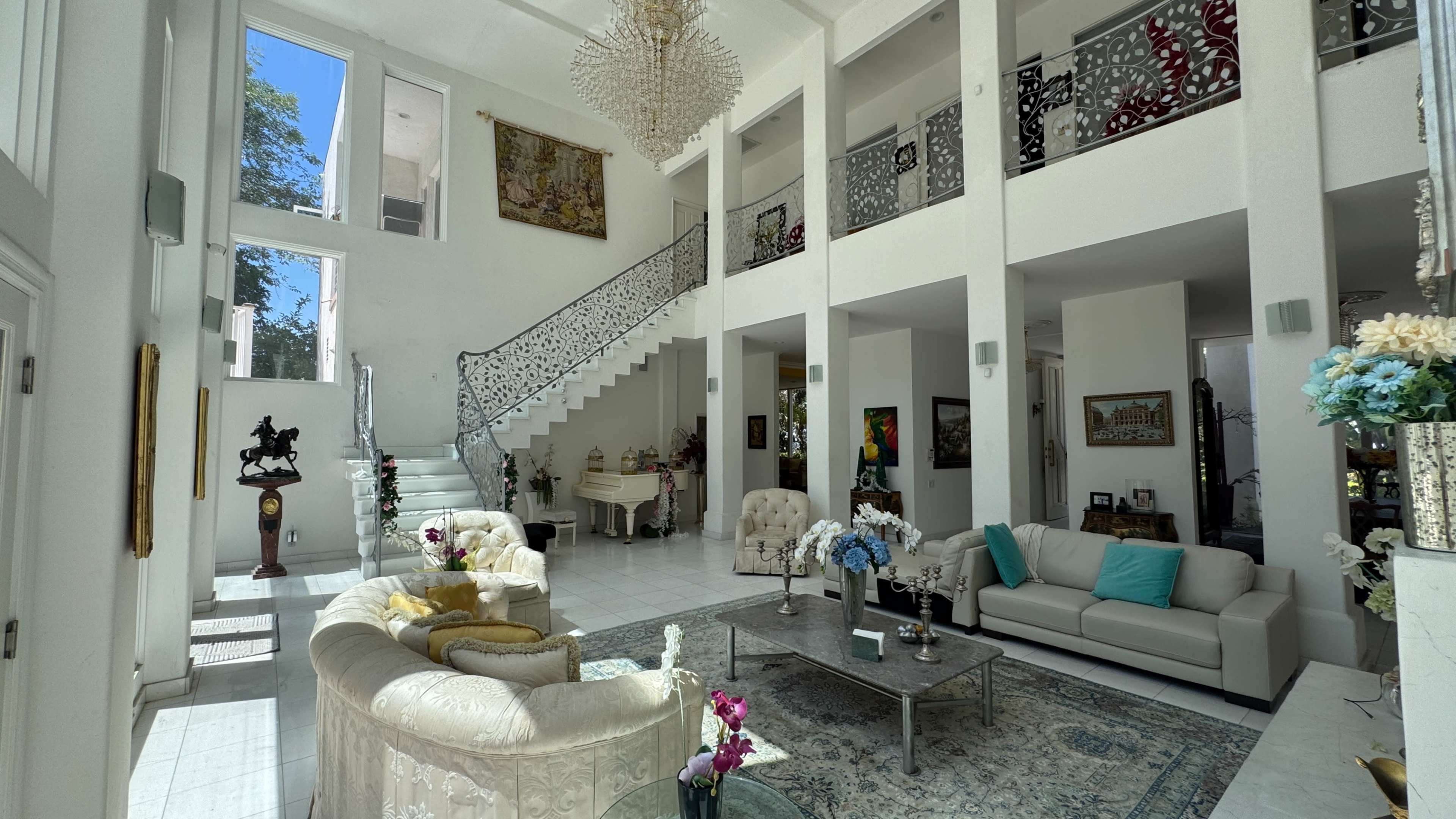The image shows a spacious, bright living room with high ceilings, featuring a central staircase, elegant furniture, and decorative artwork.