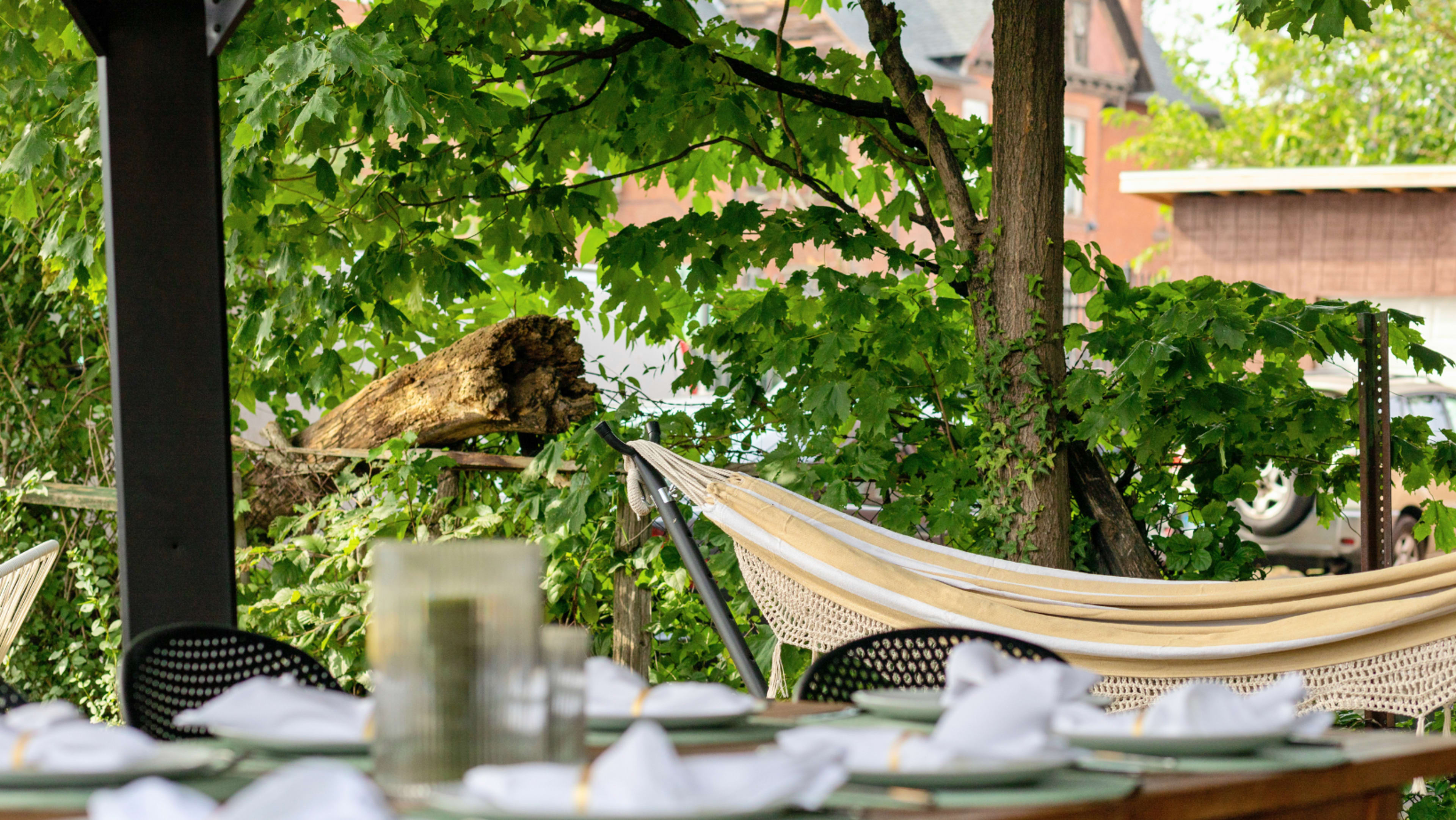 A dining table is set with white napkins in front of a hammock suspended between trees, surrounded by lush green foliage.