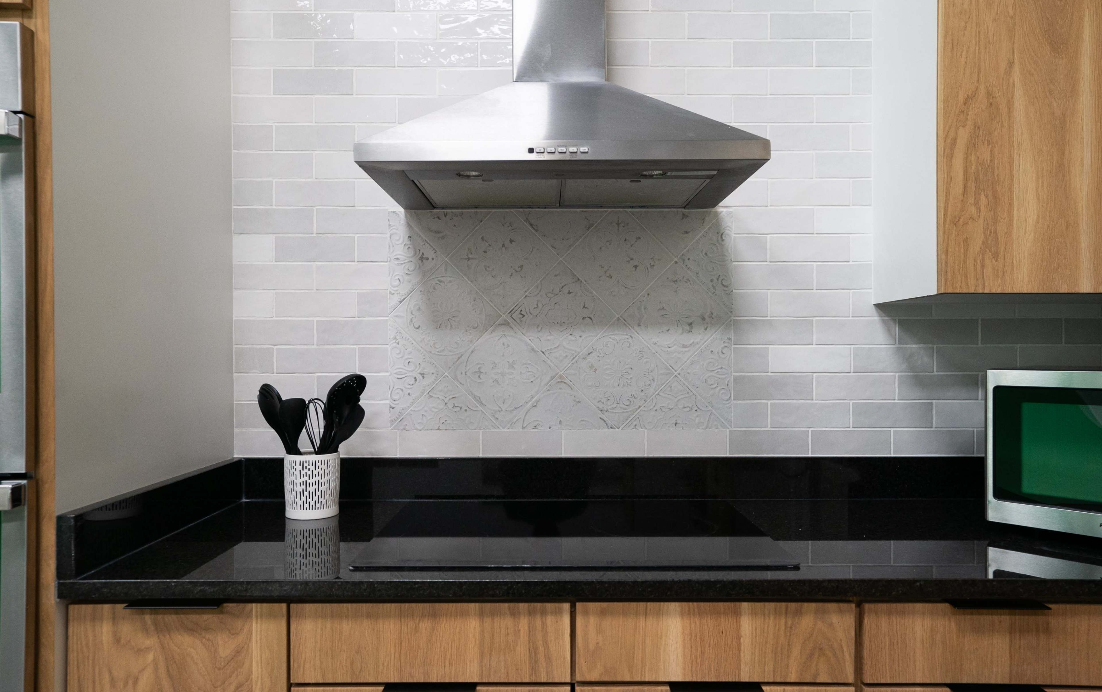 The image shows a modern kitchen with a stainless steel range hood above a black countertop and wooden cabinetry, featuring a decorative tile backsplash and a utensil holder.