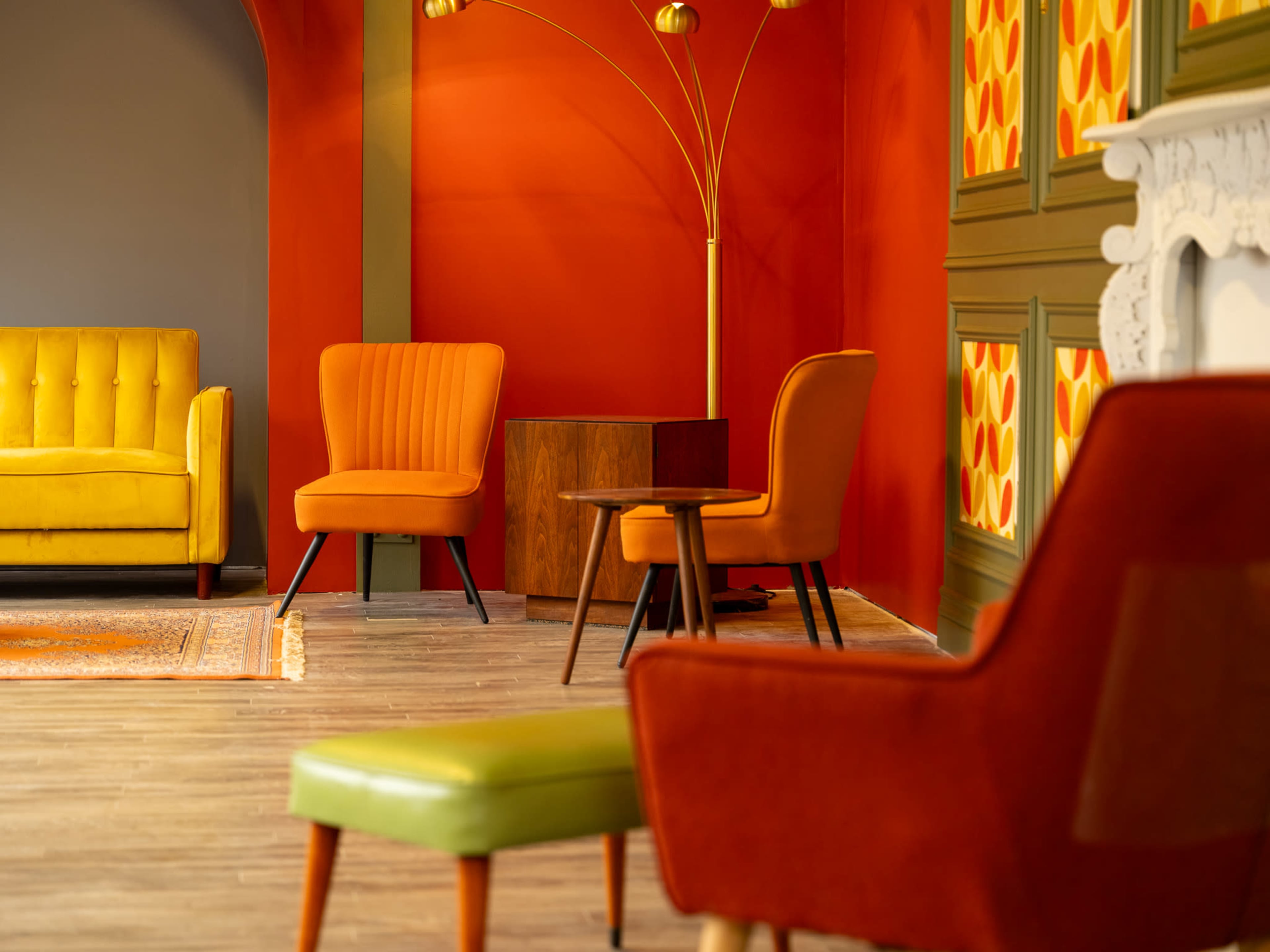 The image features a modern interior with orange and yellow furniture, including a yellow sofa and shaped armchairs, against a vibrant red and green backdrop.