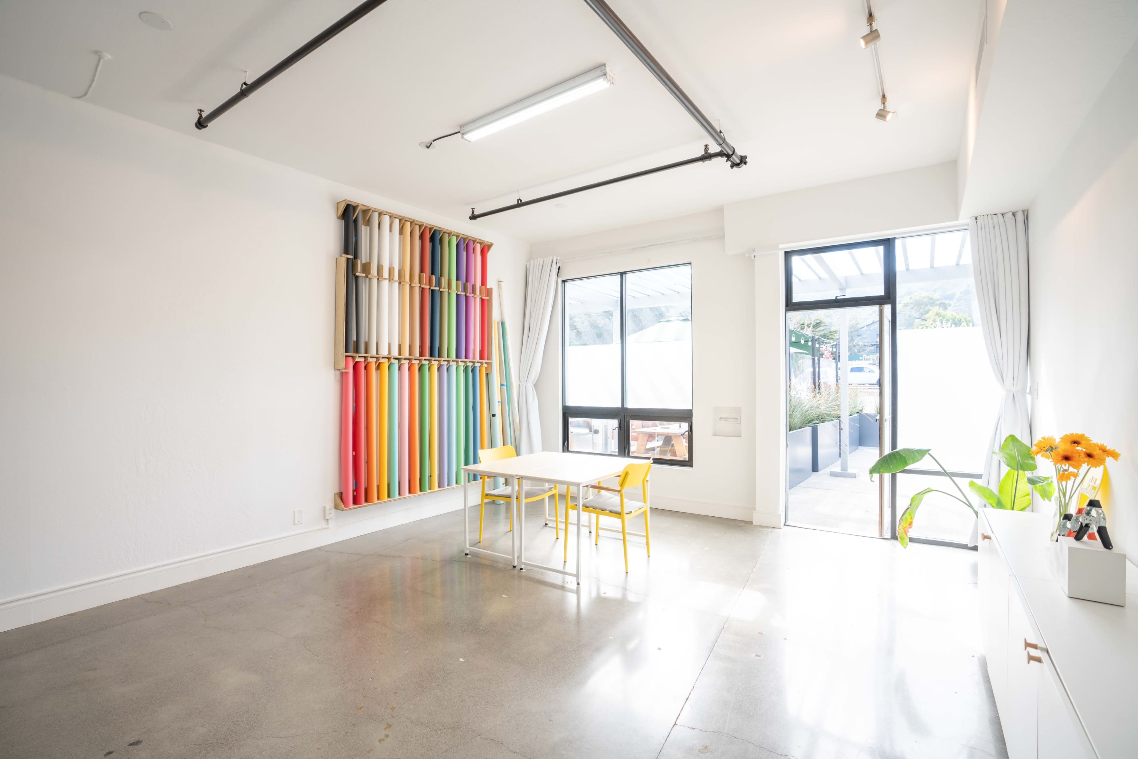 A minimalistic room with colorful fabric rolls on a wall, a white table, and two yellow chairs.