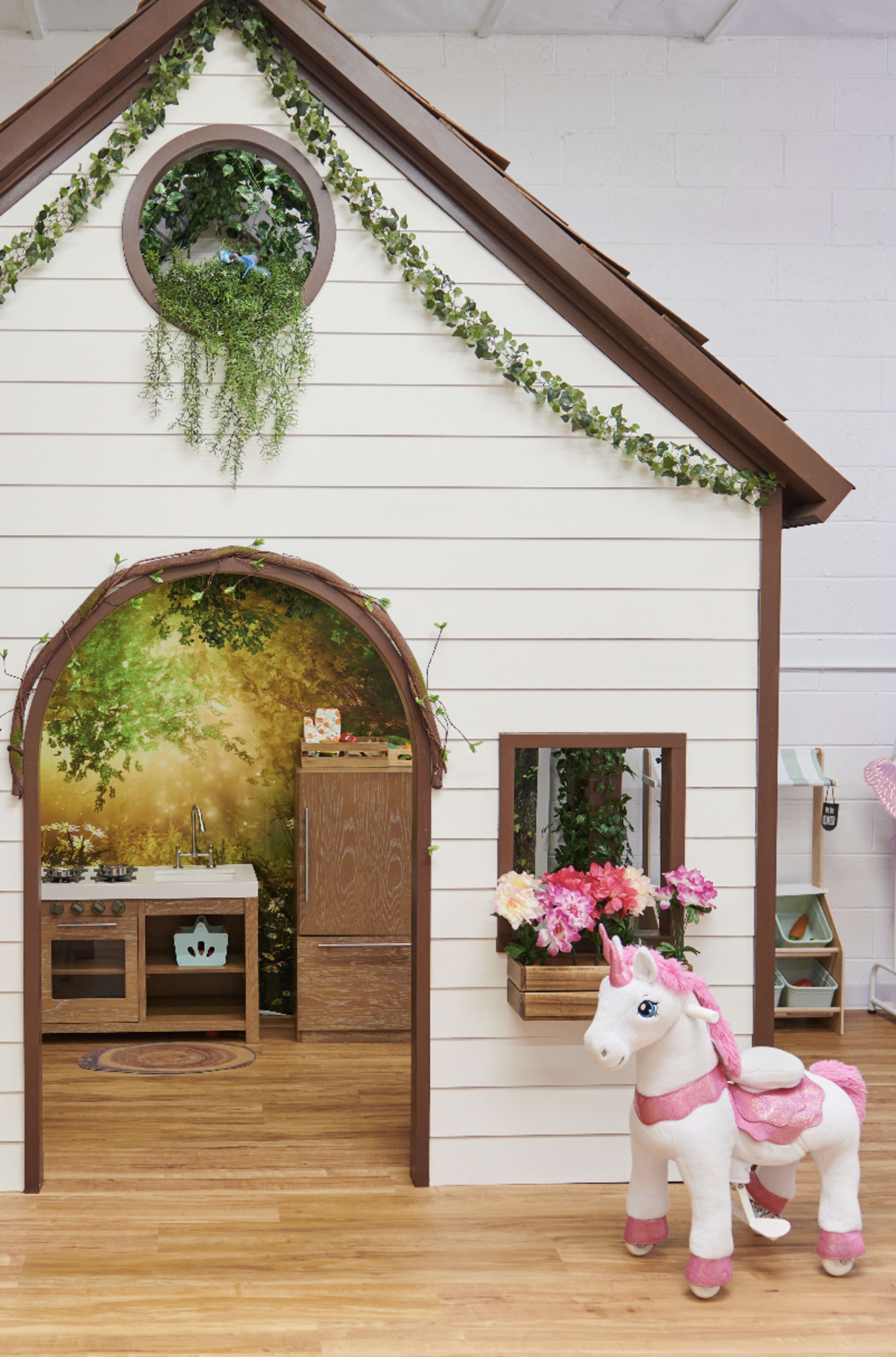 A small playhouse with a wooden archway features a kitchenette inside, while a stuffed unicorn stands outside beside a window adorned with flowers.