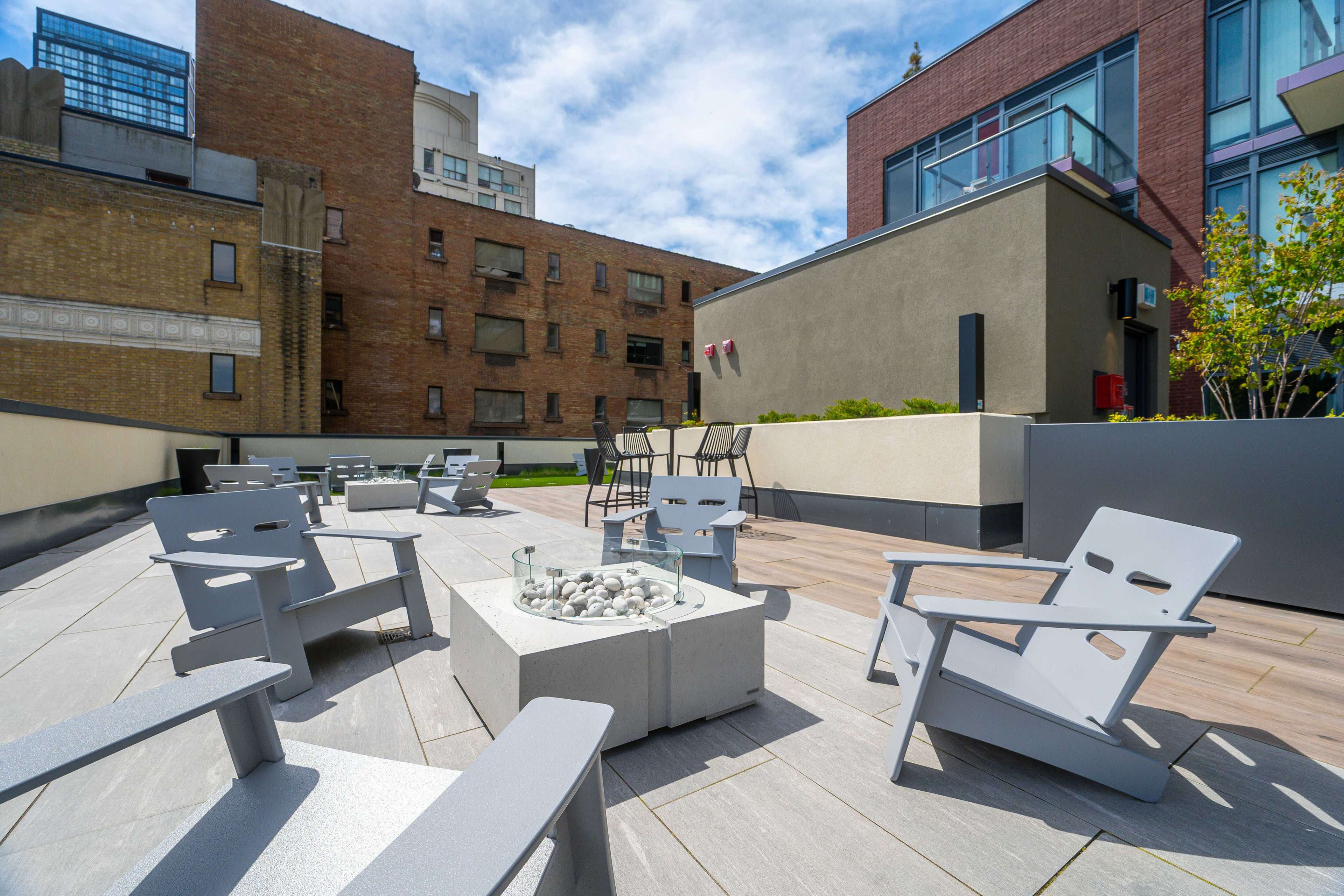 The image shows a rooftop terrace featuring modern gray chairs arranged around a circular fire pit, with a backdrop of urban buildings and scattered greenery.