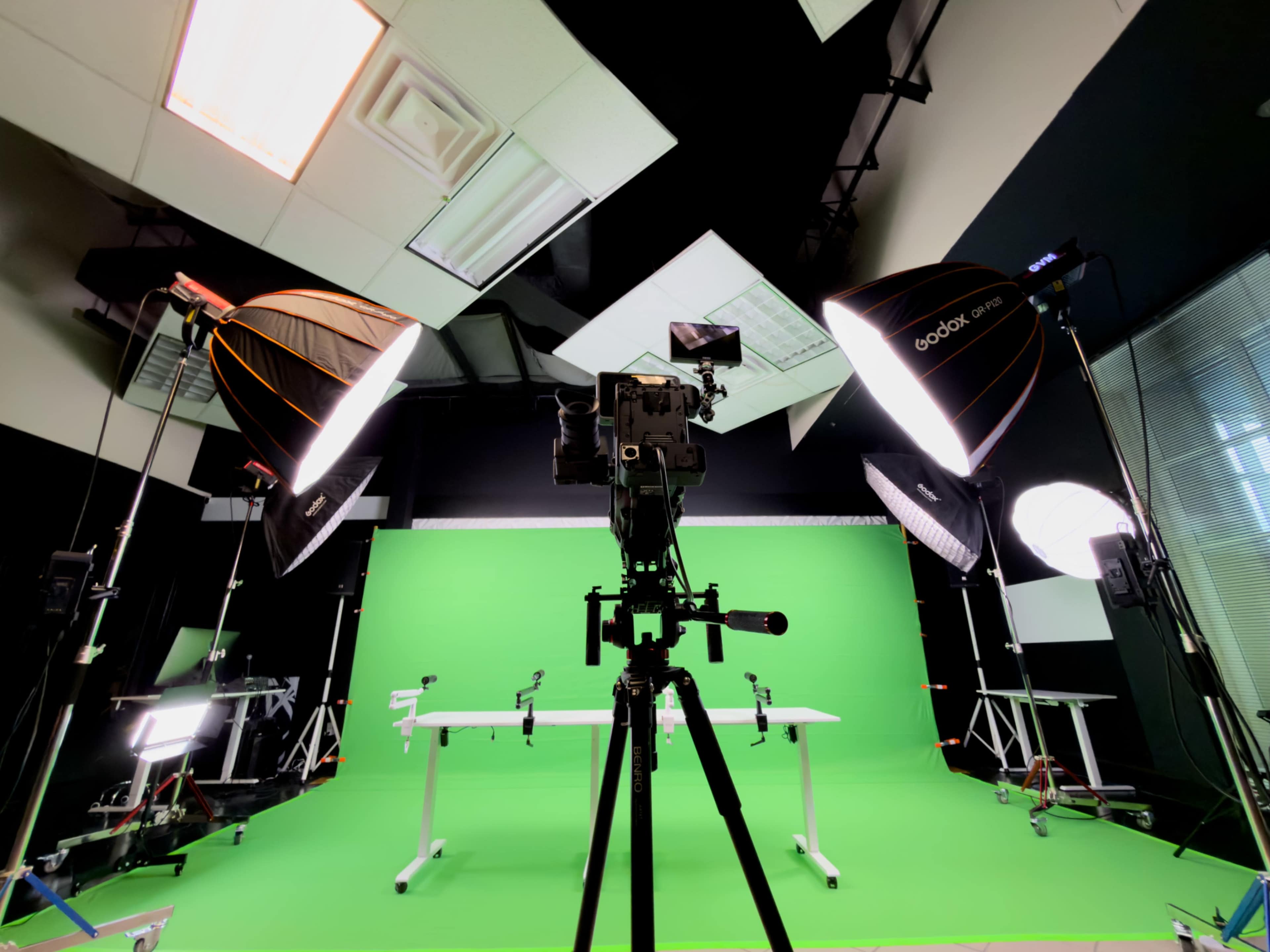 The image shows a video production setup featuring a camera on a tripod facing a green screen, with multiple lighting equipment arranged around the scene.