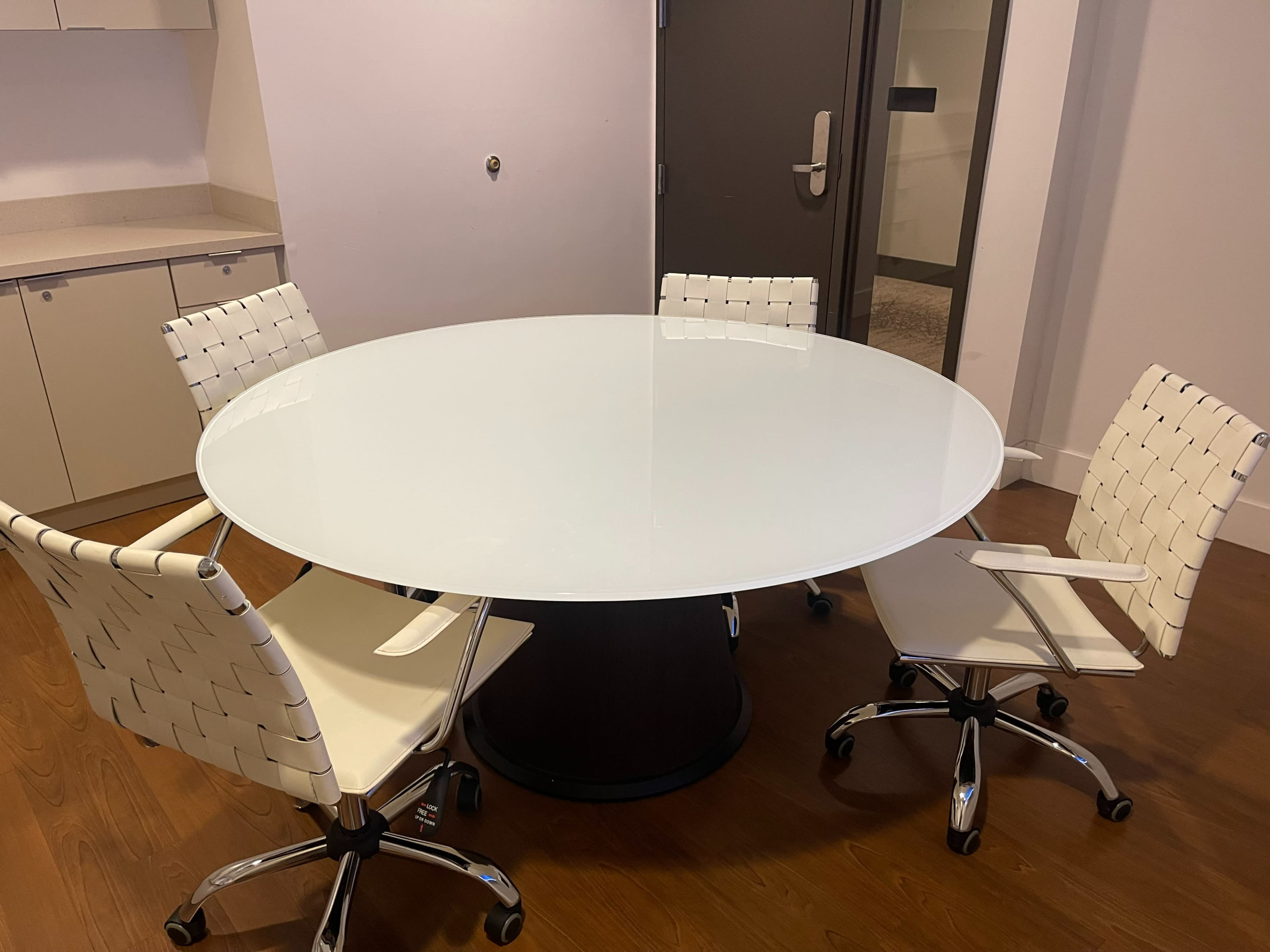 A round glass table is surrounded by four white office chairs with wheels.